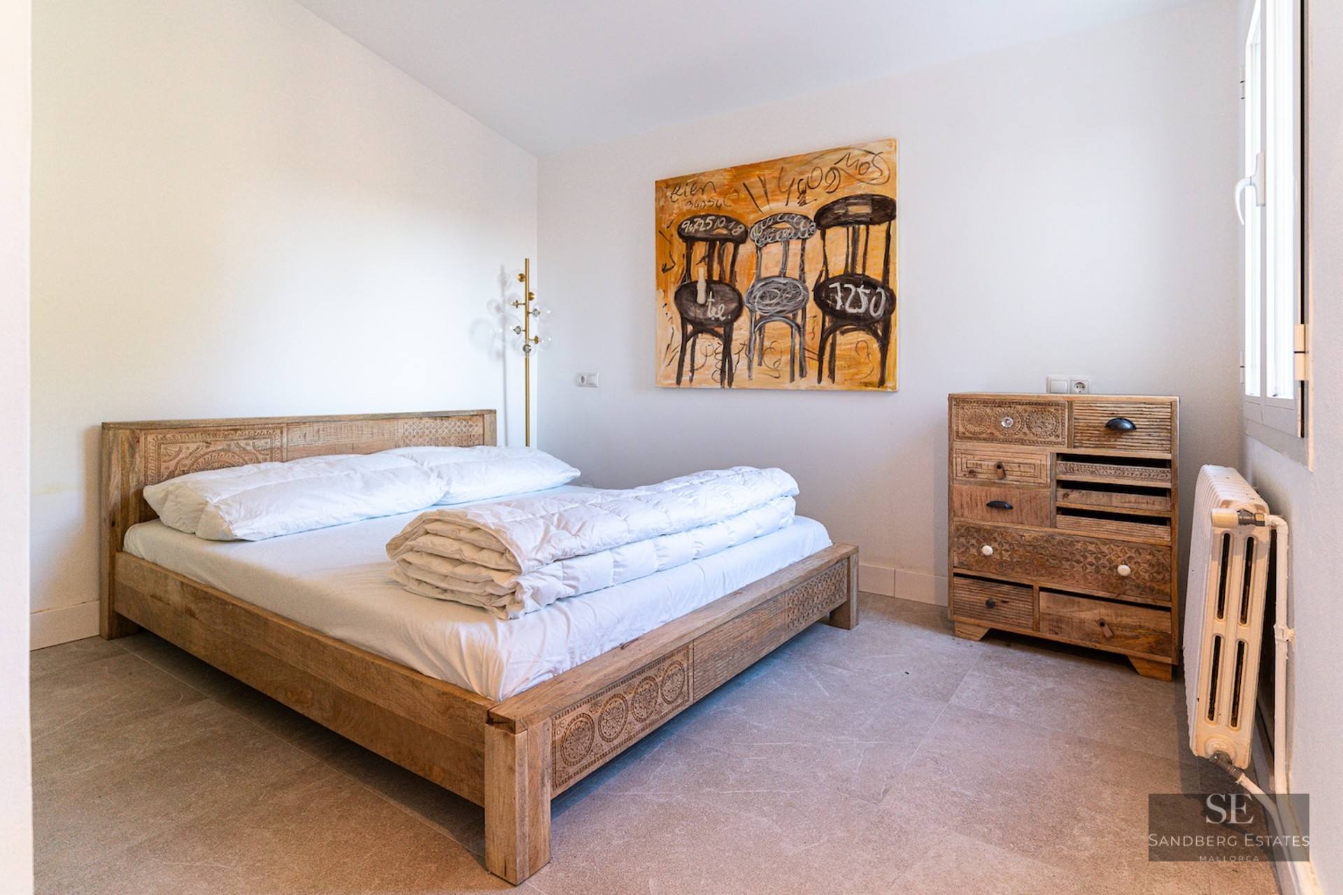Bright bedroom with a carved wooden bed, matching dresser, large abstract chair painting, and natural stone floors.