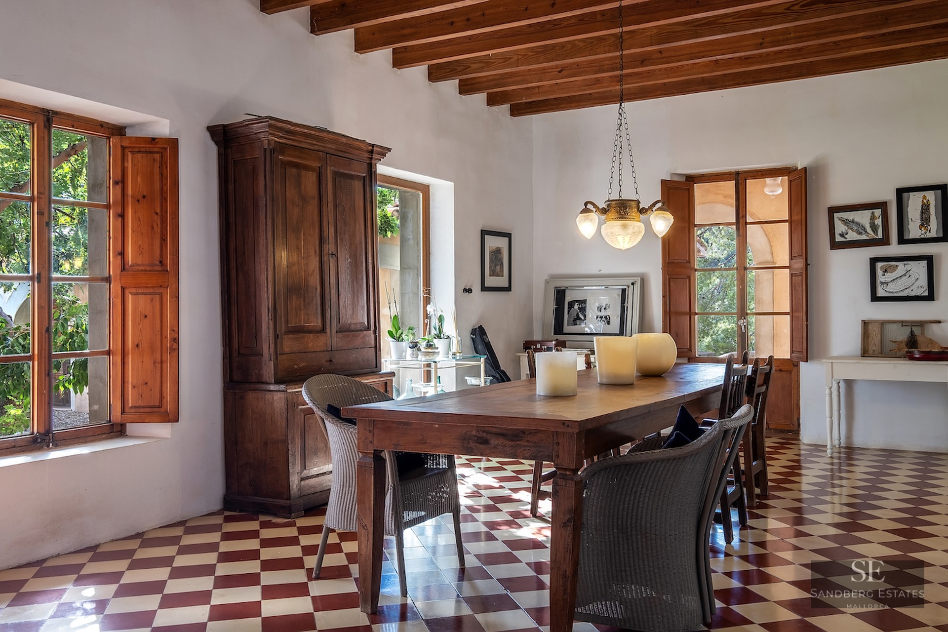 A rustic dining room with a large wood table, red and white checkered tile floor, and exposed ceiling beams.