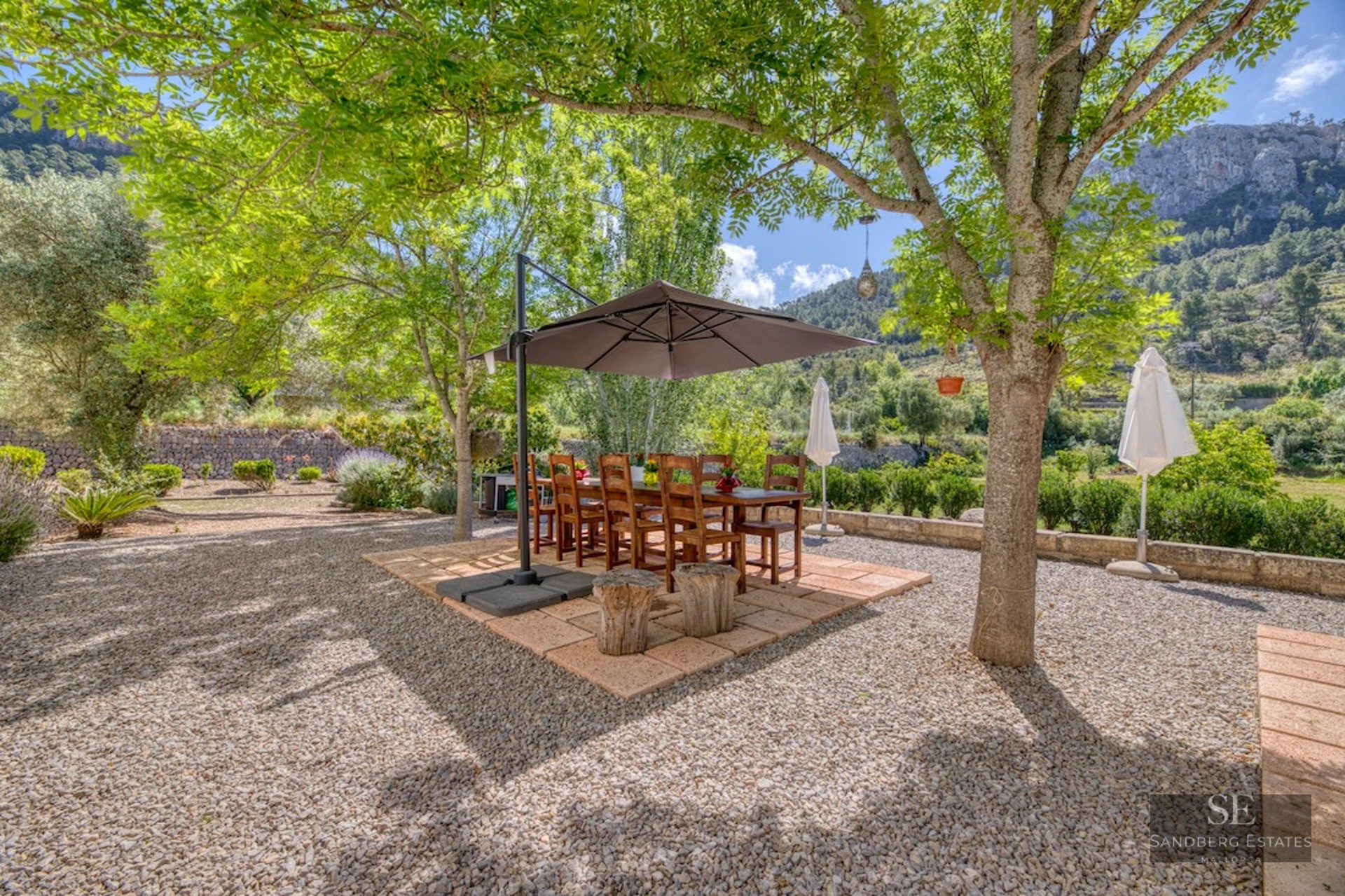 Wooden dining table and chairs under a parasol on a gravel terrace surrounded by trees and mountains.