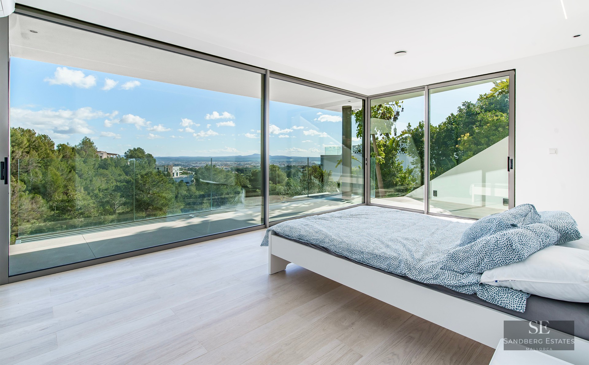 Modern bedroom with white bed and large floor-to-ceiling glass walls overlooking a lush green forest and distant city.