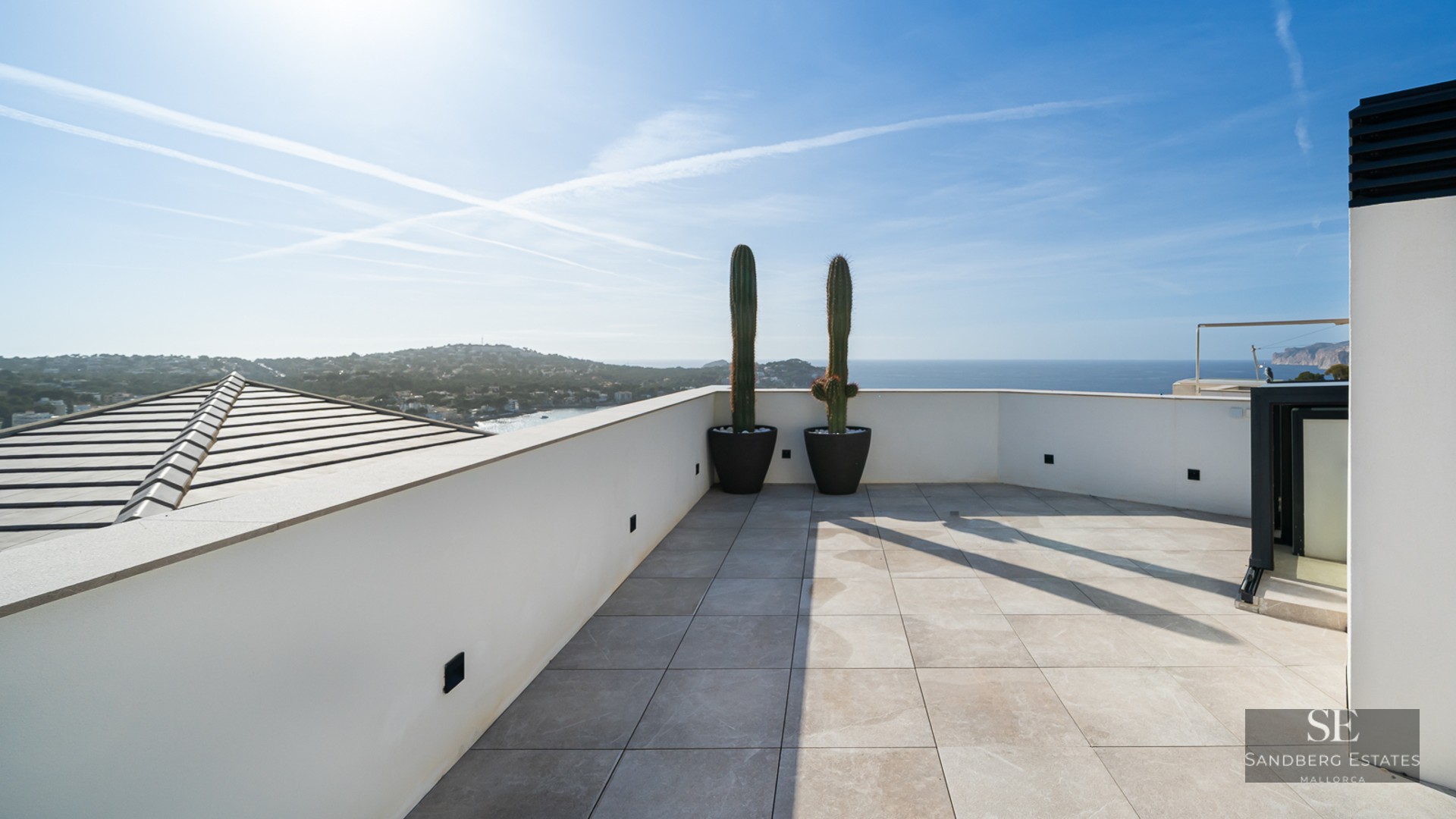 Sun-drenched modern terrace with stone tiles, white walls, and two tall cacti overlooking the Mediterranean coast.