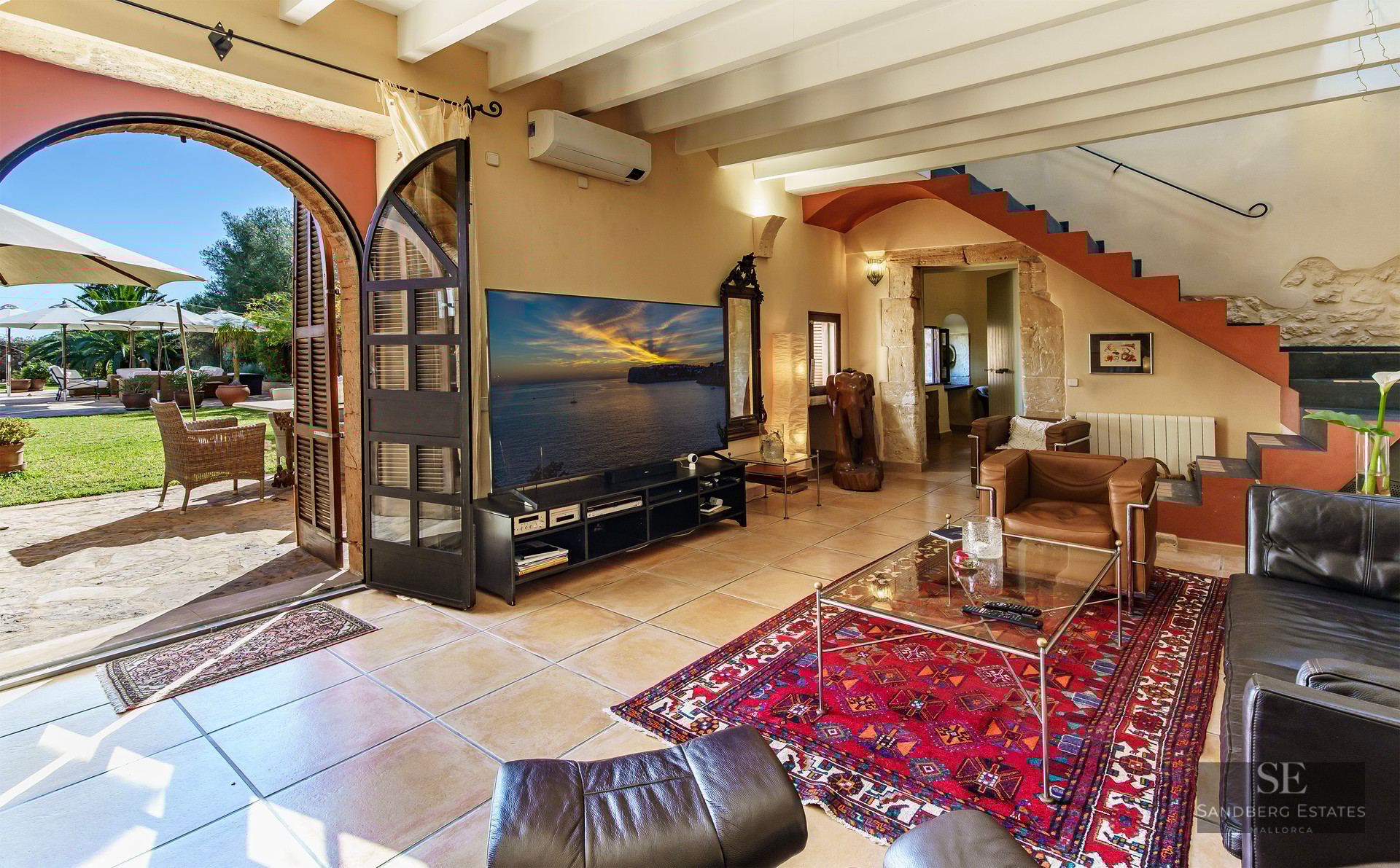 Living room with leather sofas, exposed beams, and a large arch opening to a sunny garden with umbrellas.