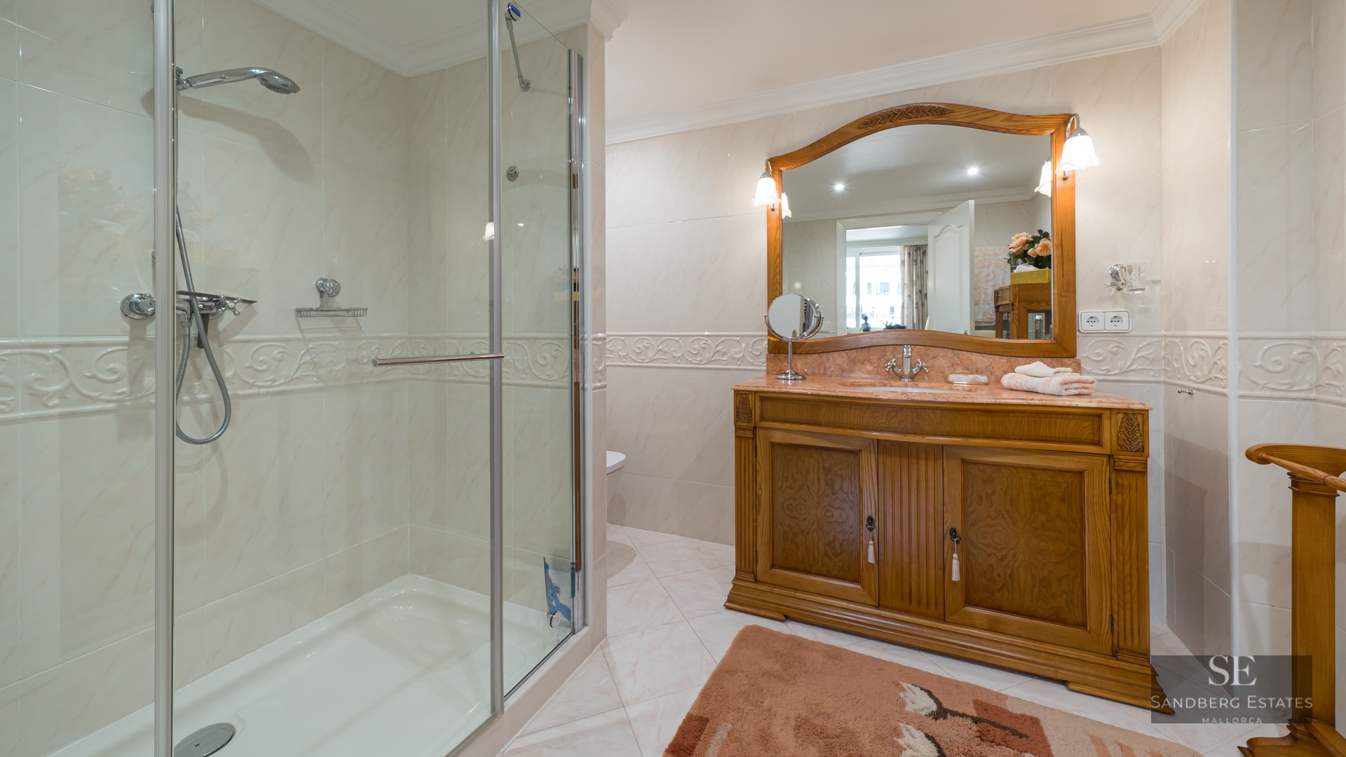 A classic bathroom featuring a glass-enclosed shower, cream marble wall tiles, and a large wooden vanity with a mirror.