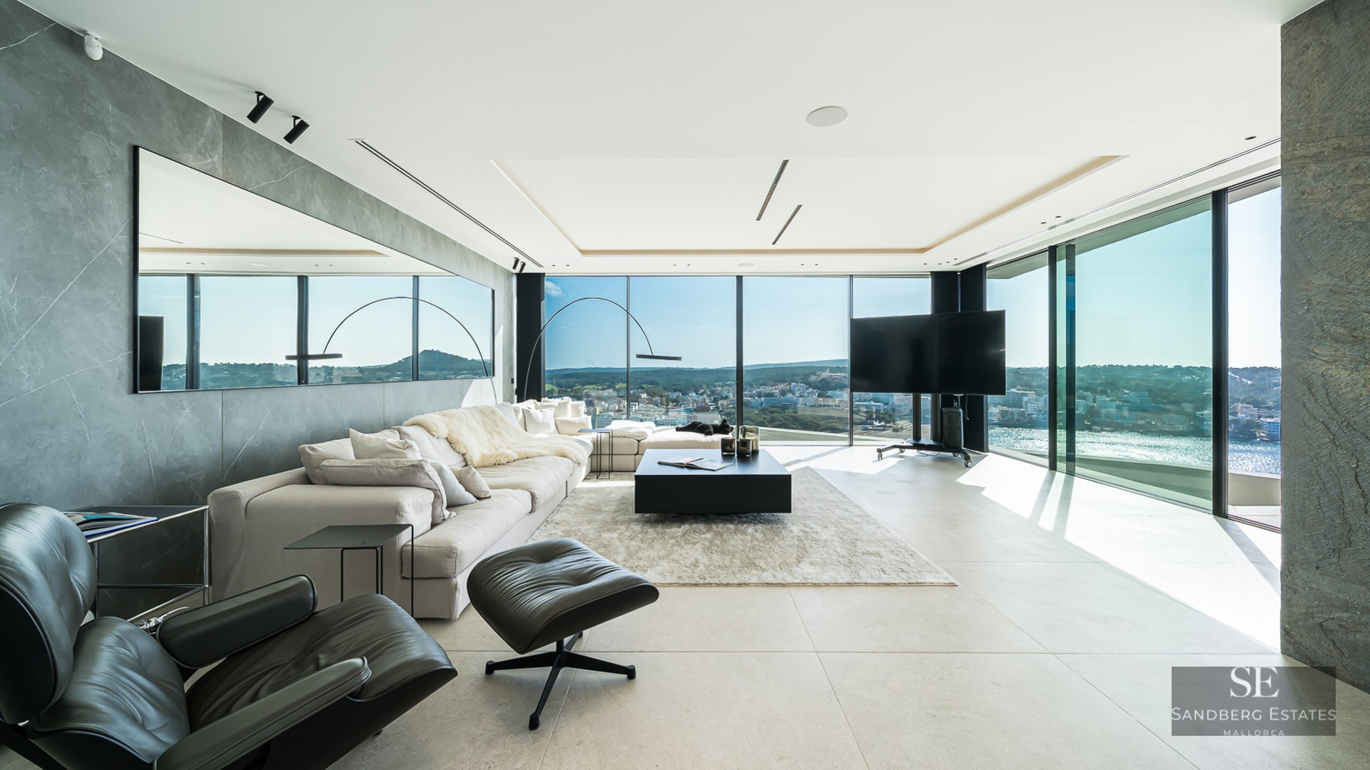 Modern living room with a large white sofa, black leather lounge chair, and floor-to-ceiling windows showing a coastal view.
