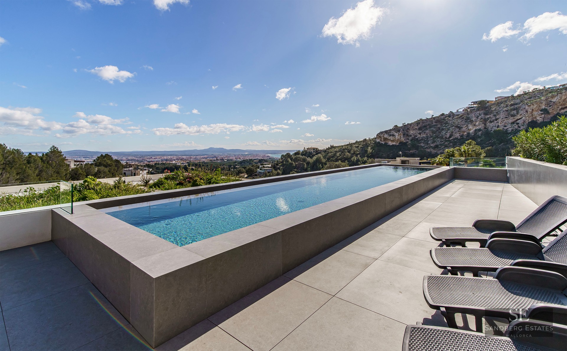 Infinity pool on a stone terrace with sun loungers overlooking a valley and city landscape.