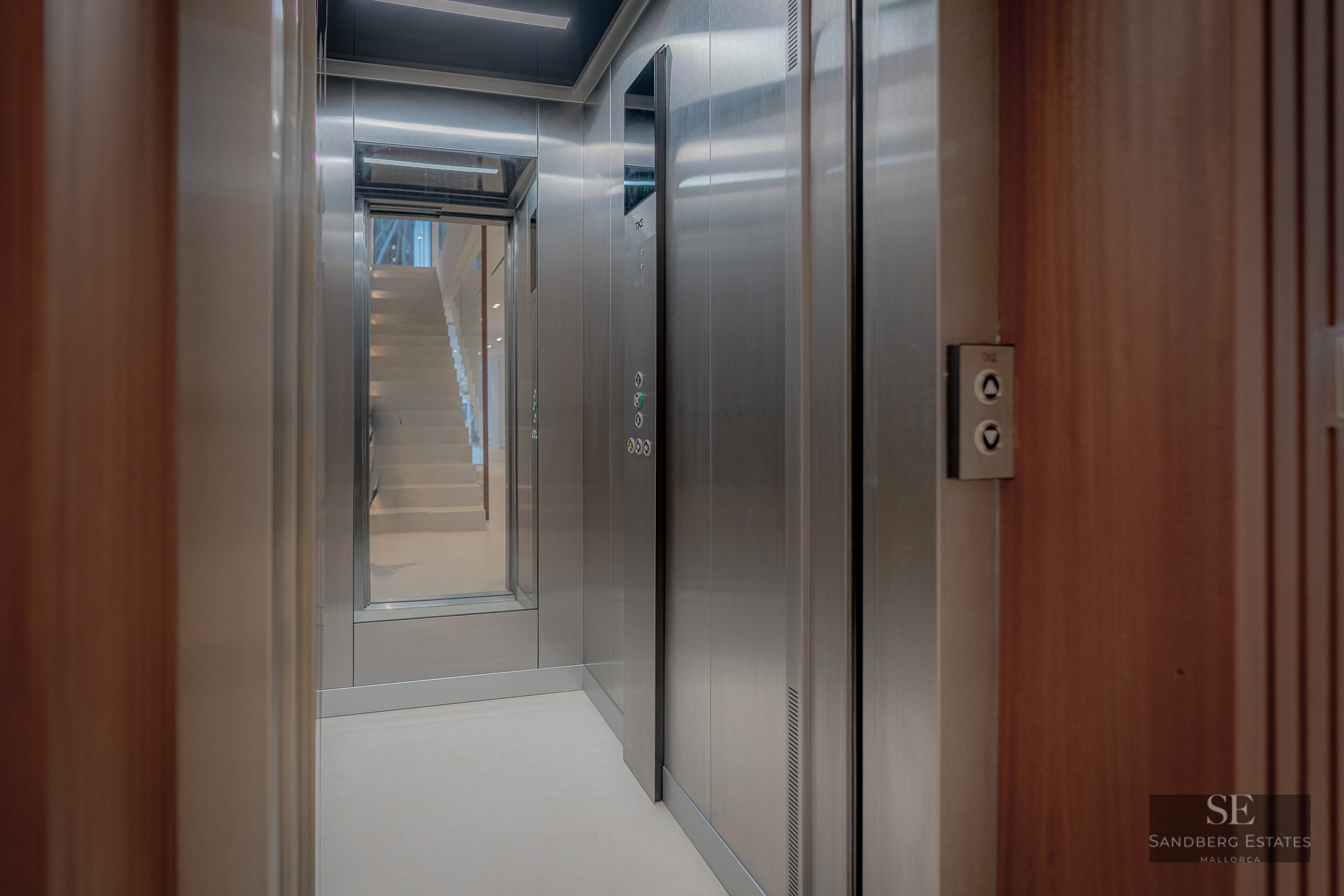 Interior of a modern elevator with stainless steel finishes, wood paneling, and a mirror reflecting a staircase.