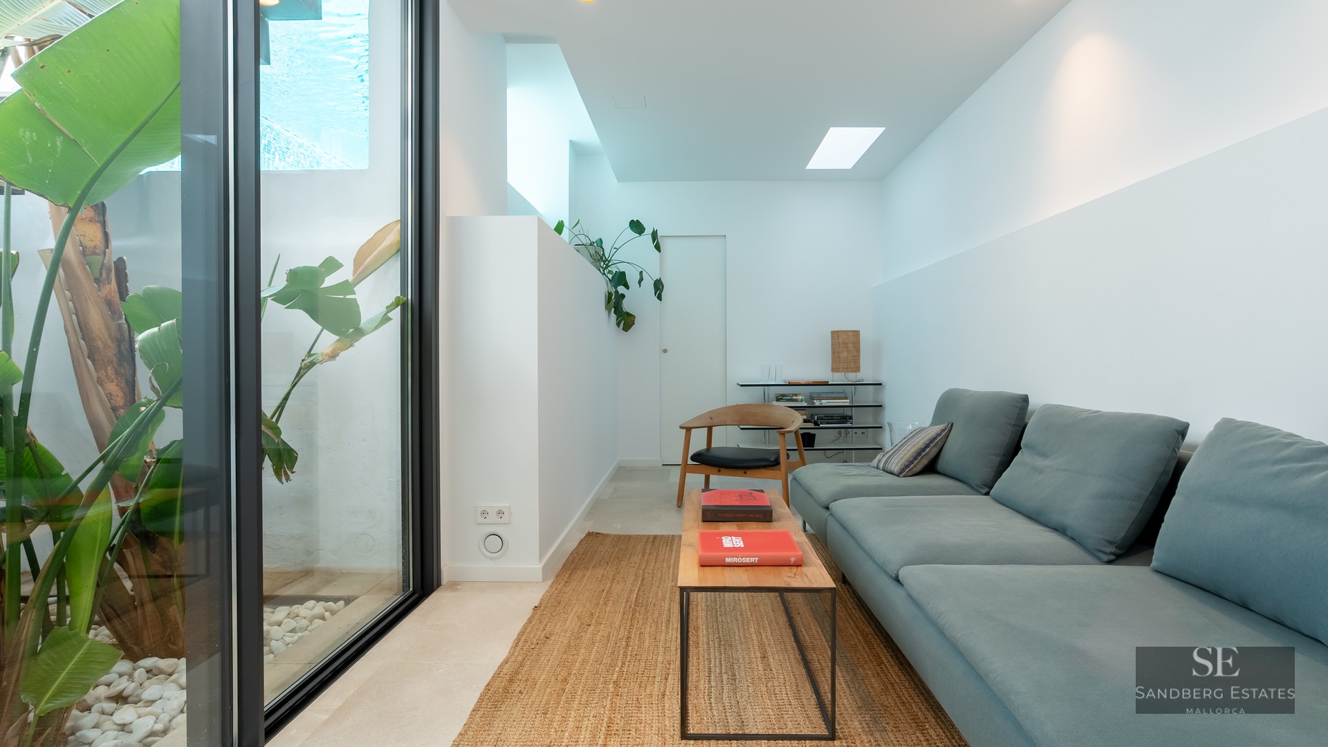Modern living room with blue sofa, wooden coffee table, jute rug, and glass wall looking out to tropical plants.