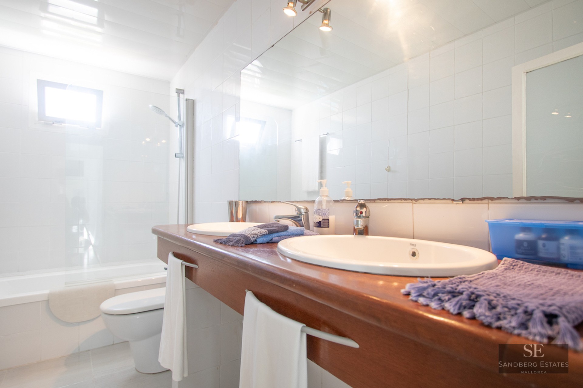 Modern white bathroom featuring a wooden double sink vanity, large mirror, and bathtub with glass screen.