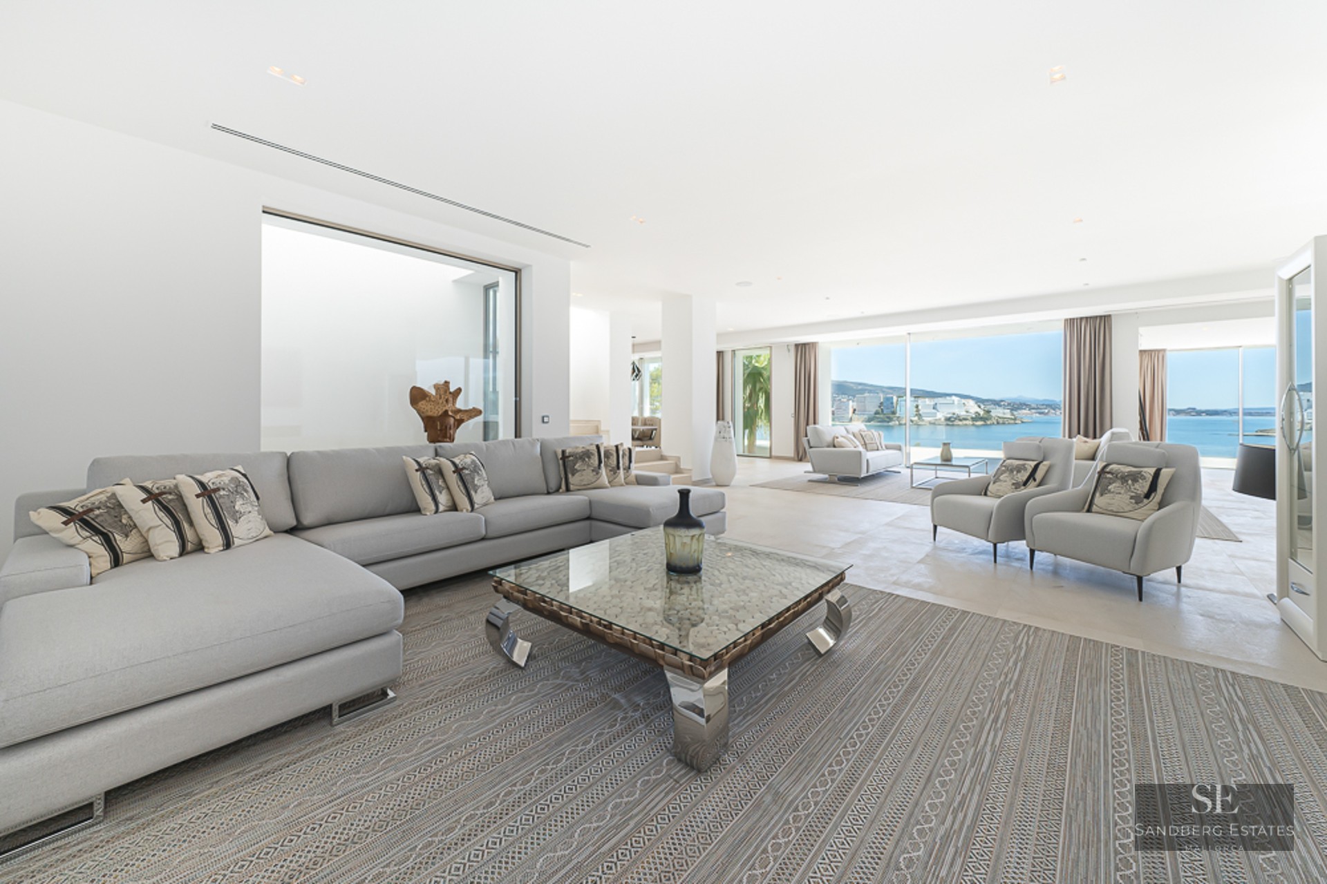 Spacious modern living room with light gray sofas, a glass coffee table, and large windows overlooking the Mediterranean sea.