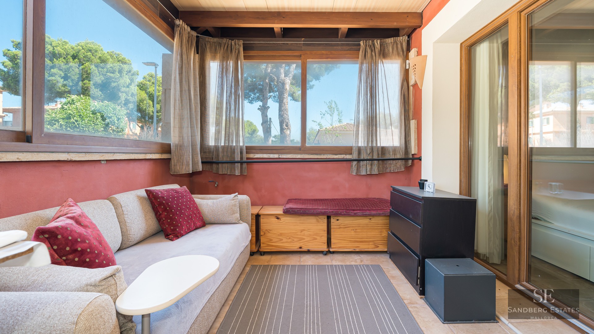 Sunlit living area with beige sofa, wooden beams, terracotta walls, and large windows overlooking green trees.