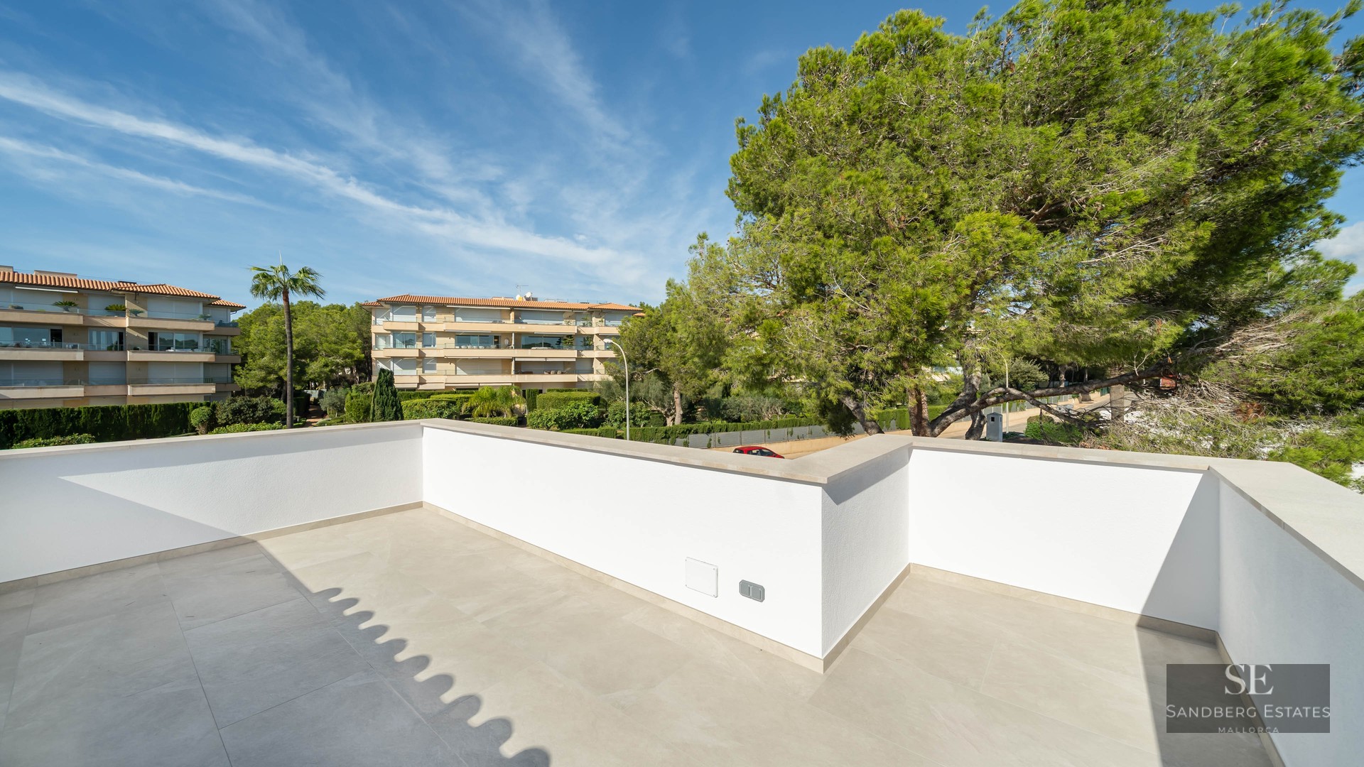 Spacious modern terrace with white walls and light stone flooring, overlooking lush pine trees and buildings.