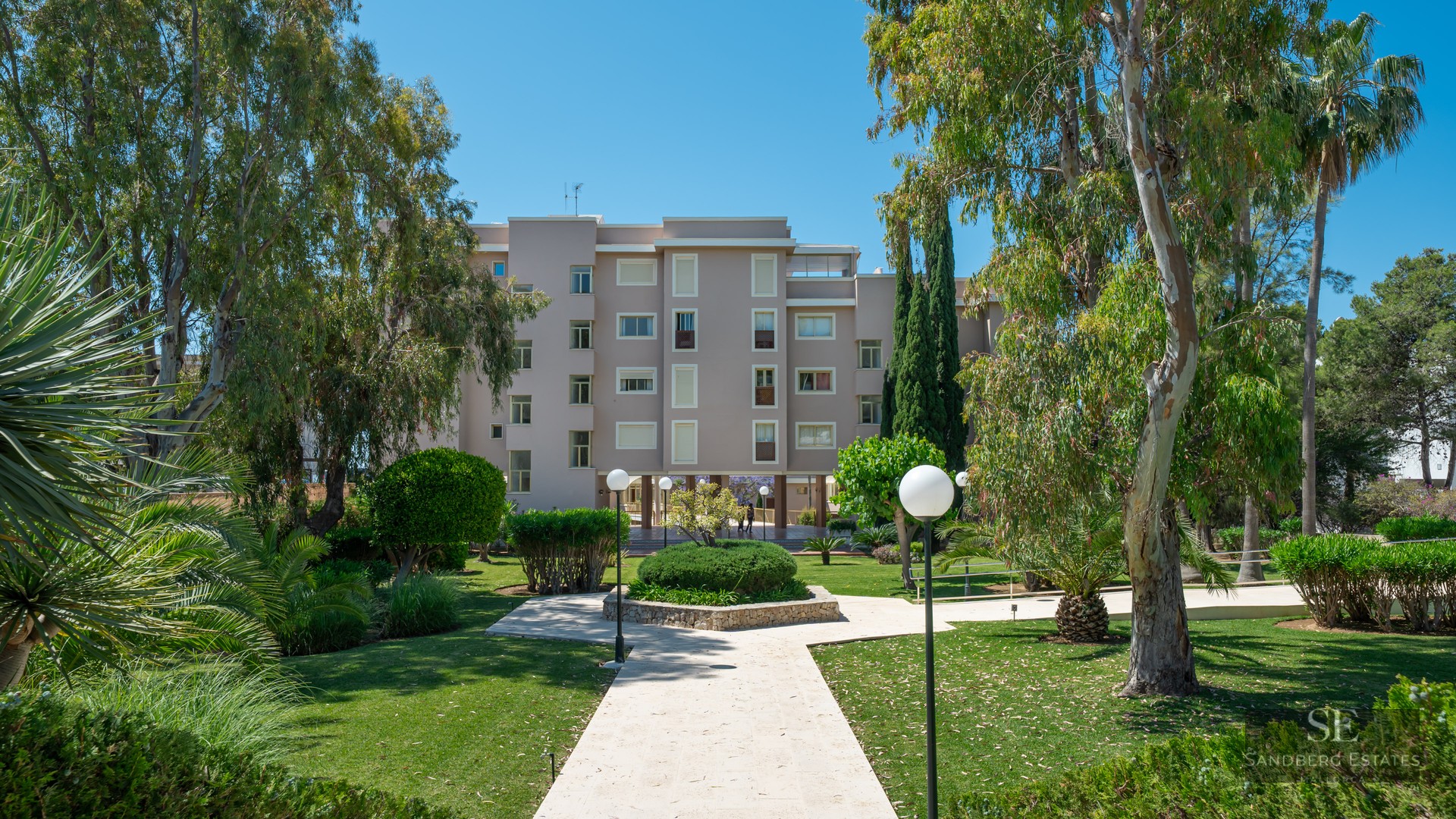 A paved pathway leads through a lush garden with trees and shrubs toward a multi-story apartment building.