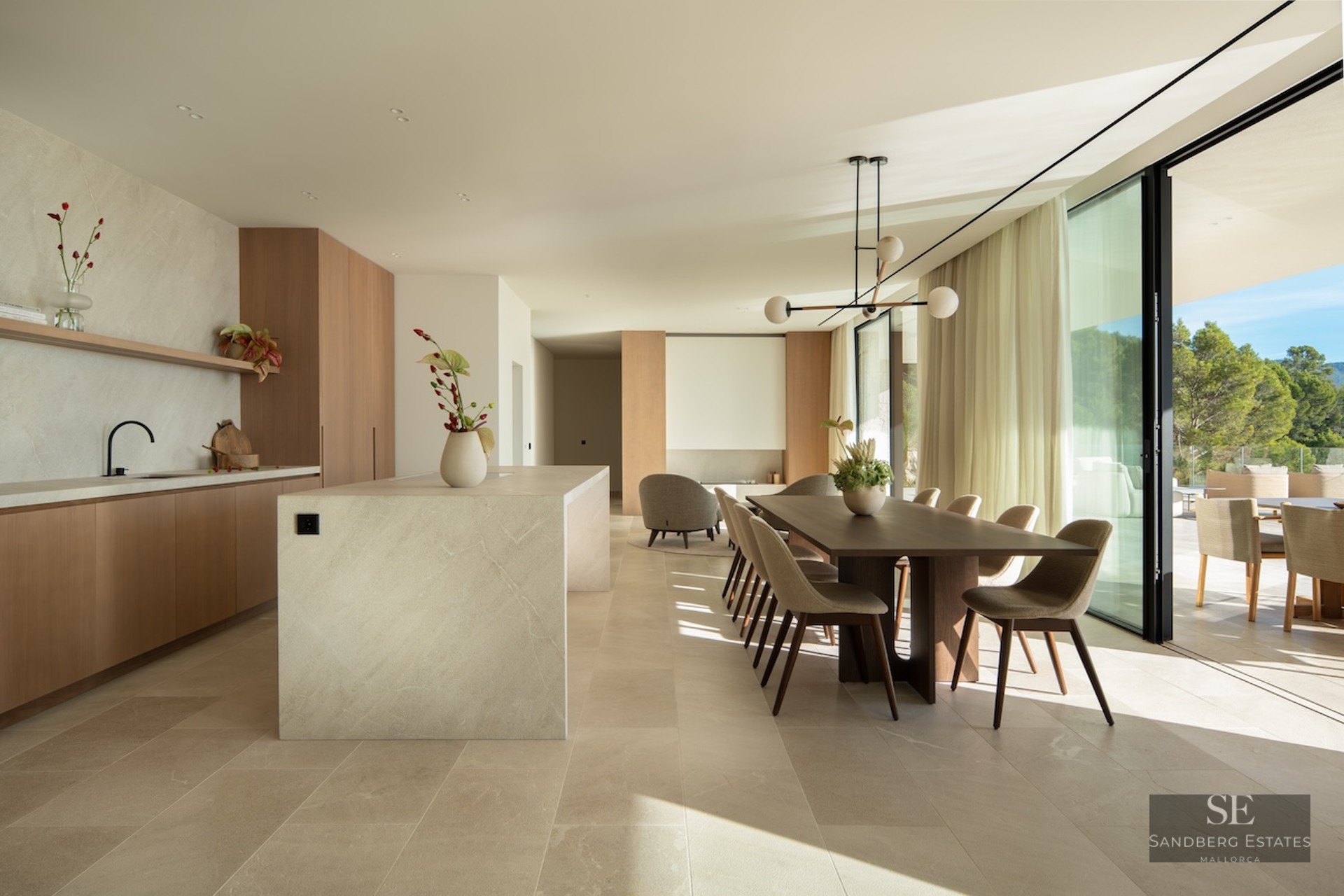 Modern dining room with a wooden table, stone kitchen island, and large floor-to-ceiling windows overlooking a forest.