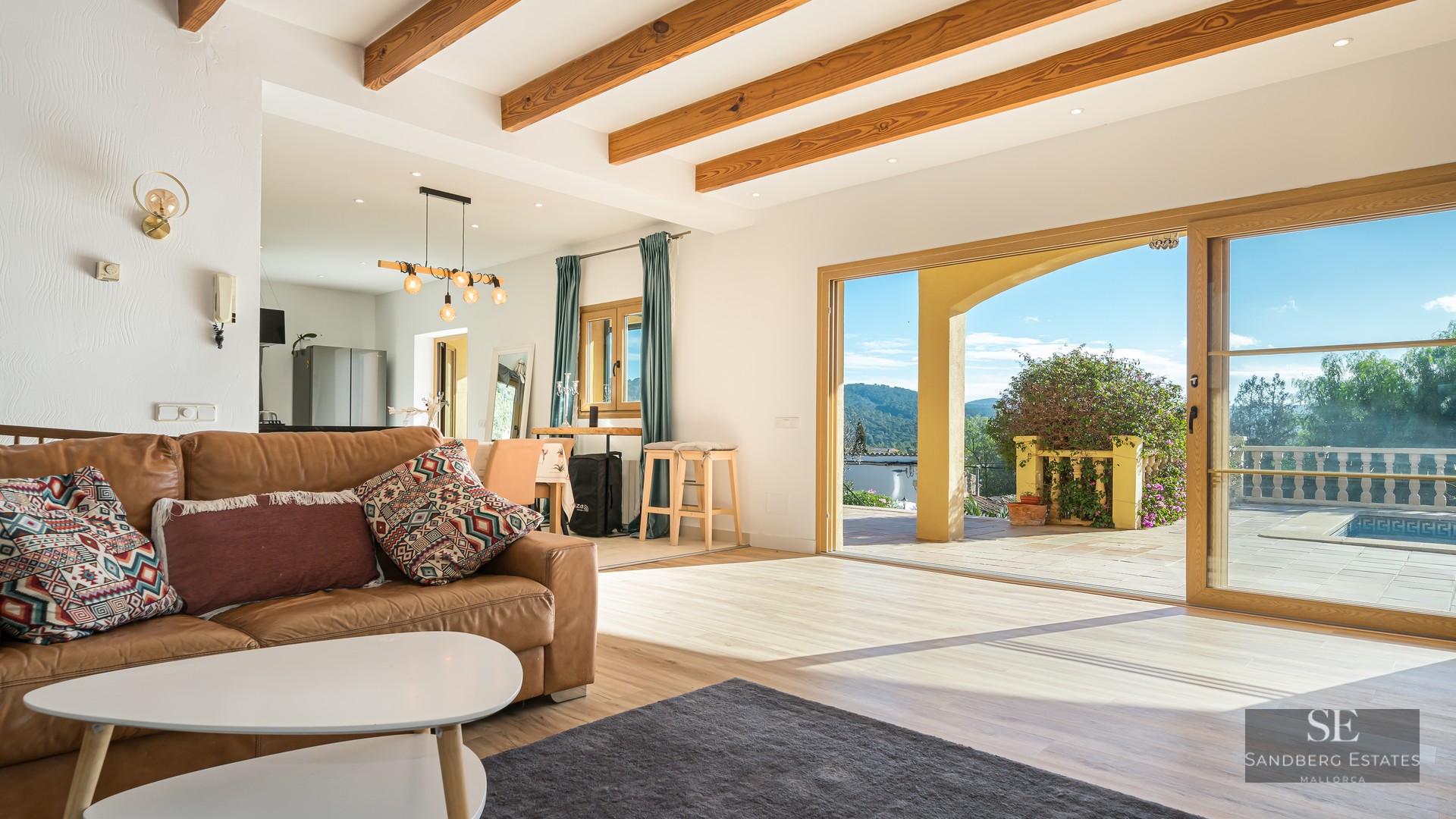 Living room with brown leather sofa, exposed wood beams, and large sliding doors opening to a terrace with mountain views.