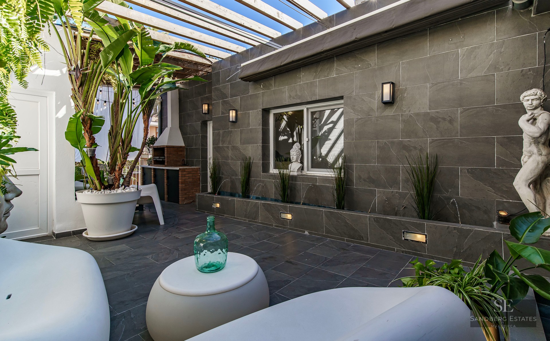 Luxury patio with grey stone walls, white modern furniture, a wooden pergola, and tropical plants.