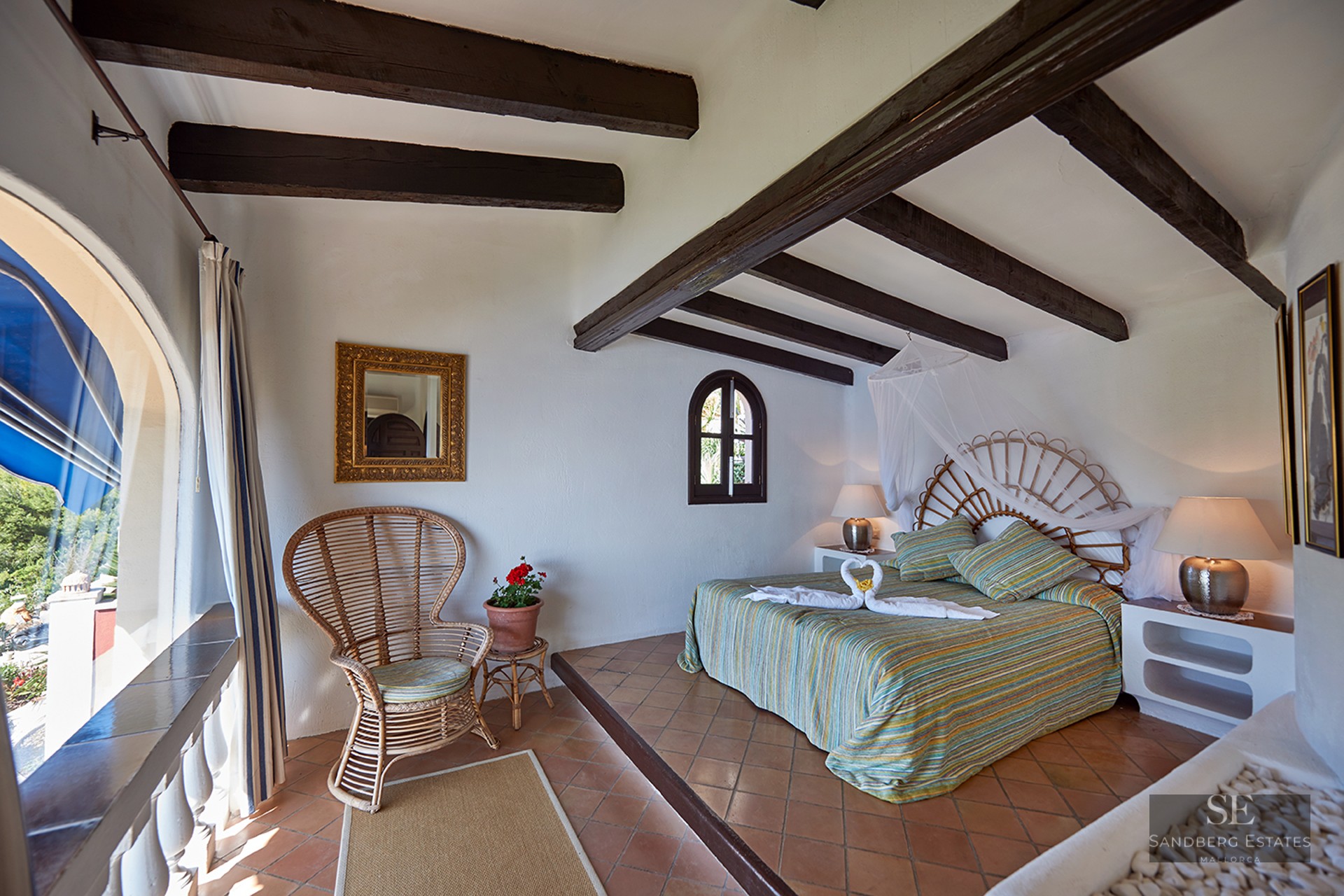 Sunlit bedroom featuring dark wood ceiling beams, a wicker headboard, terracotta floors, and an arched balcony window.