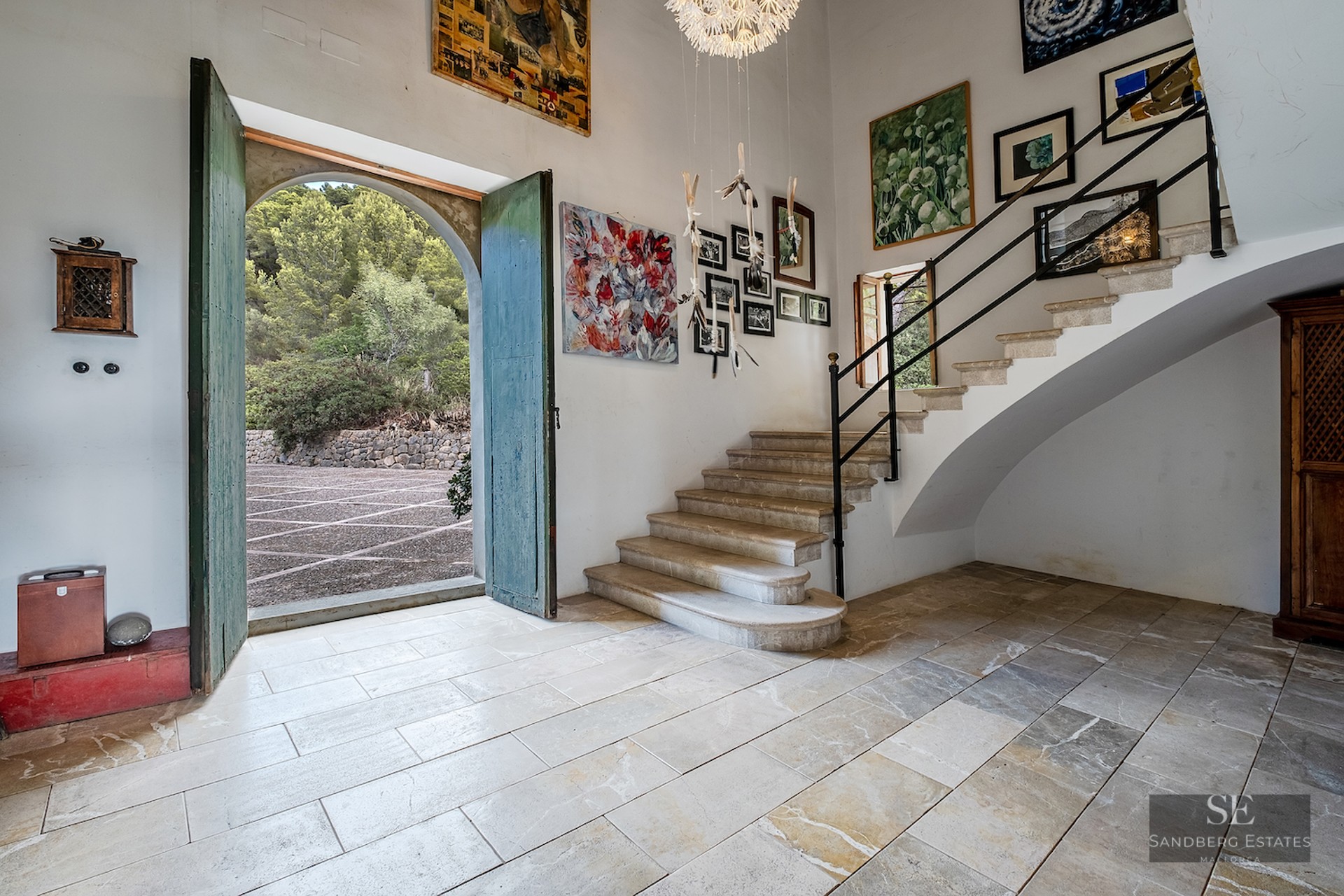 Grand entrance hall with stone floors, high ceilings, a stone staircase, and large green arched doors open to the outside.