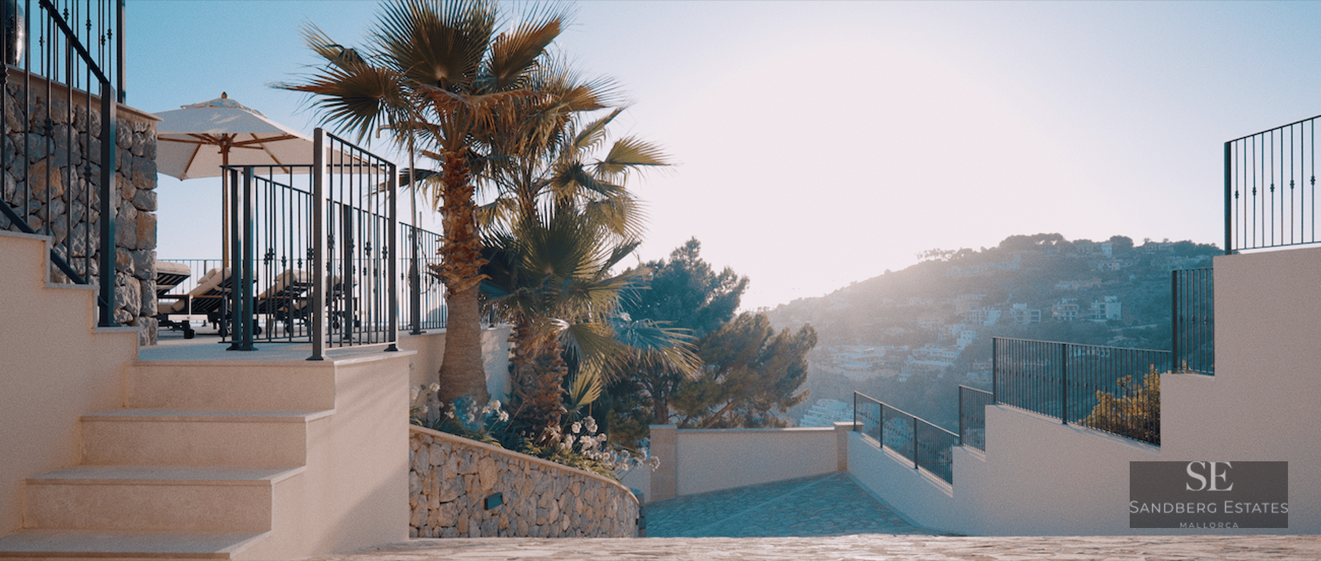 Stone stairs leading to a sunlit terrace with palm trees and lounge chairs overlooking a mountain valley.