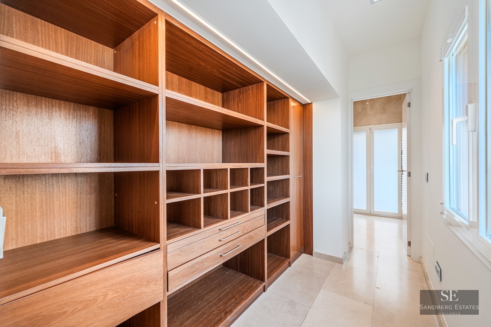 Modern walk-in closet with warm wood shelving, marble floors, and integrated LED lighting next to a bright window.