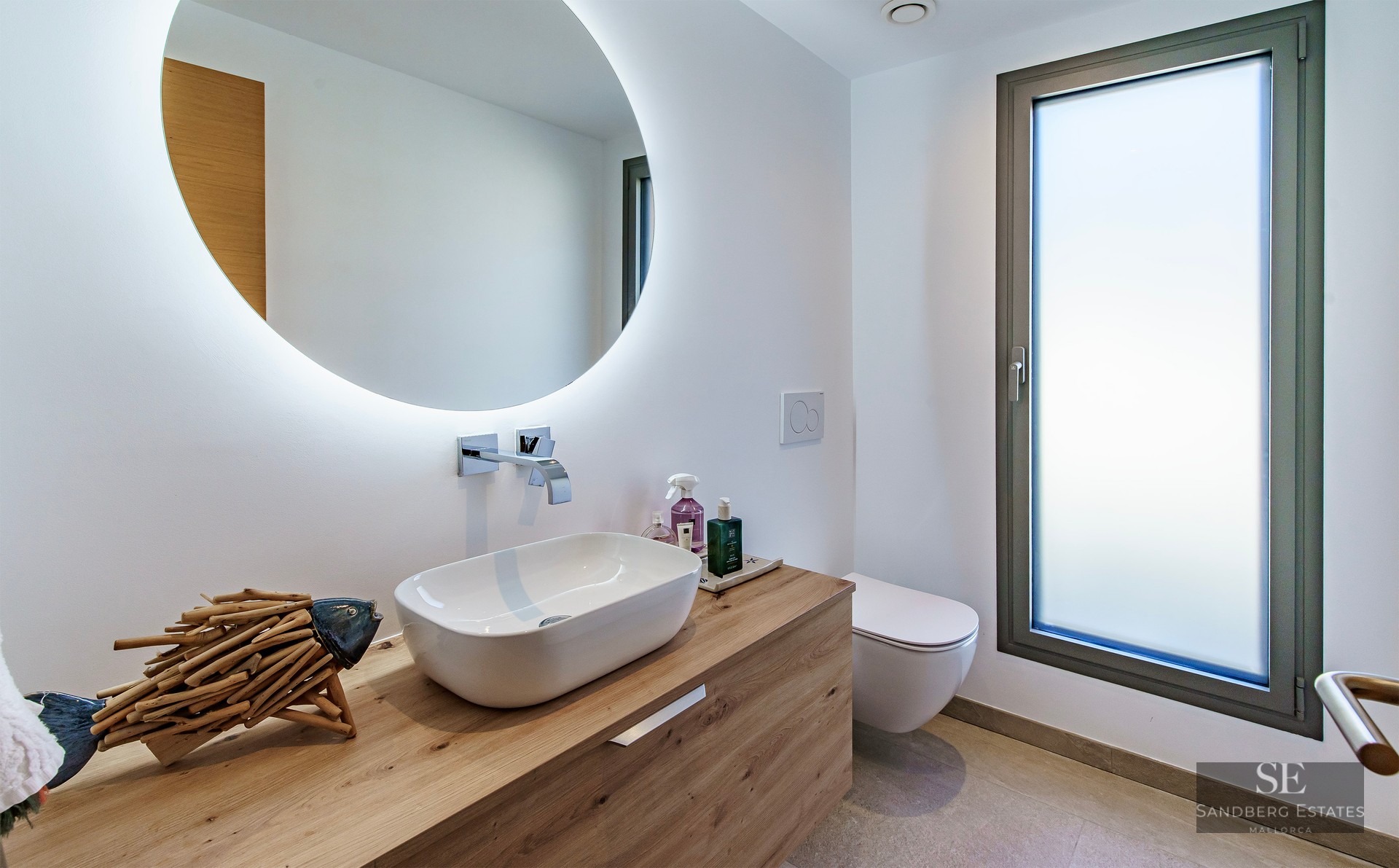 Modern bathroom featuring a white vessel sink on a wooden vanity, a large round backlit mirror, and a frosted glass window.