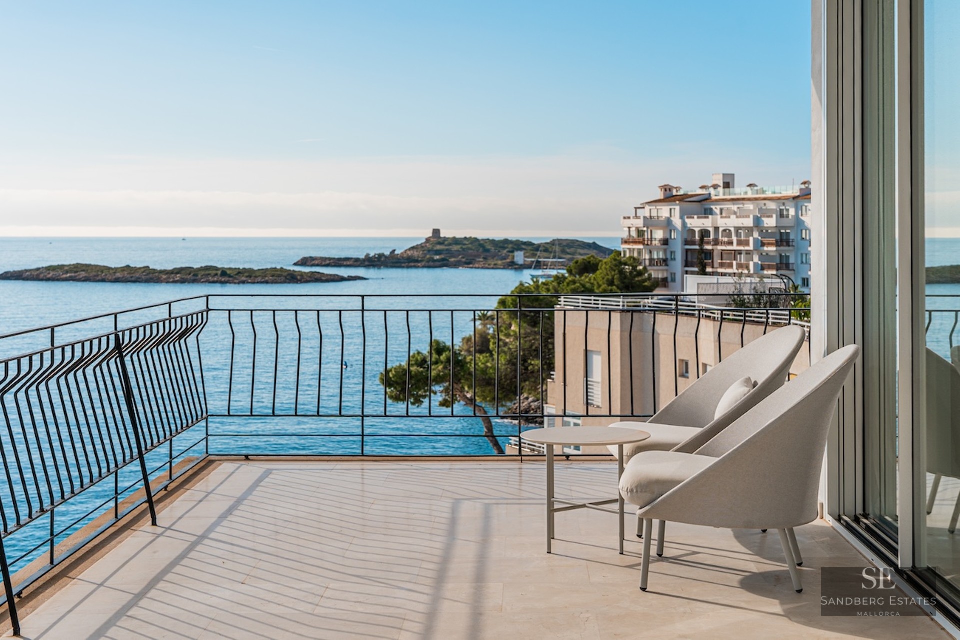 Modern balcony with grey armchairs overlooking a sparkling blue sea with islands and coastal buildings.