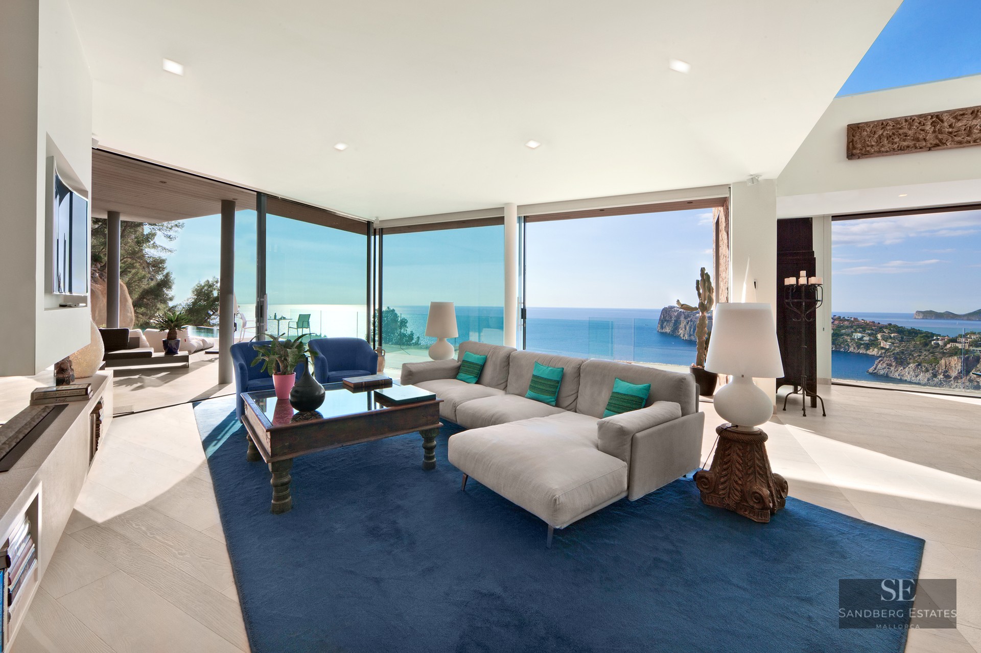 Modern living room featuring a gray sectional sofa, large blue rug, and floor-to-ceiling windows overlooking the ocean.