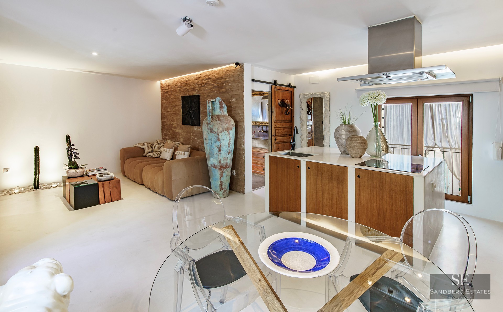 Open-plan living room and kitchen featuring a brown sofa against a brick wall, a large turquoise vase, and glass table.
