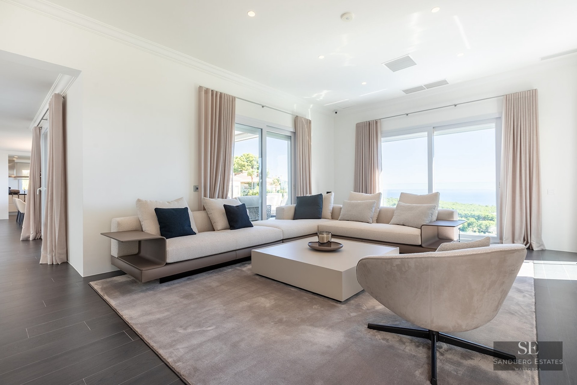 Spacious white living room with beige sectional sofa, designer armchair, and large windows overlooking the ocean.