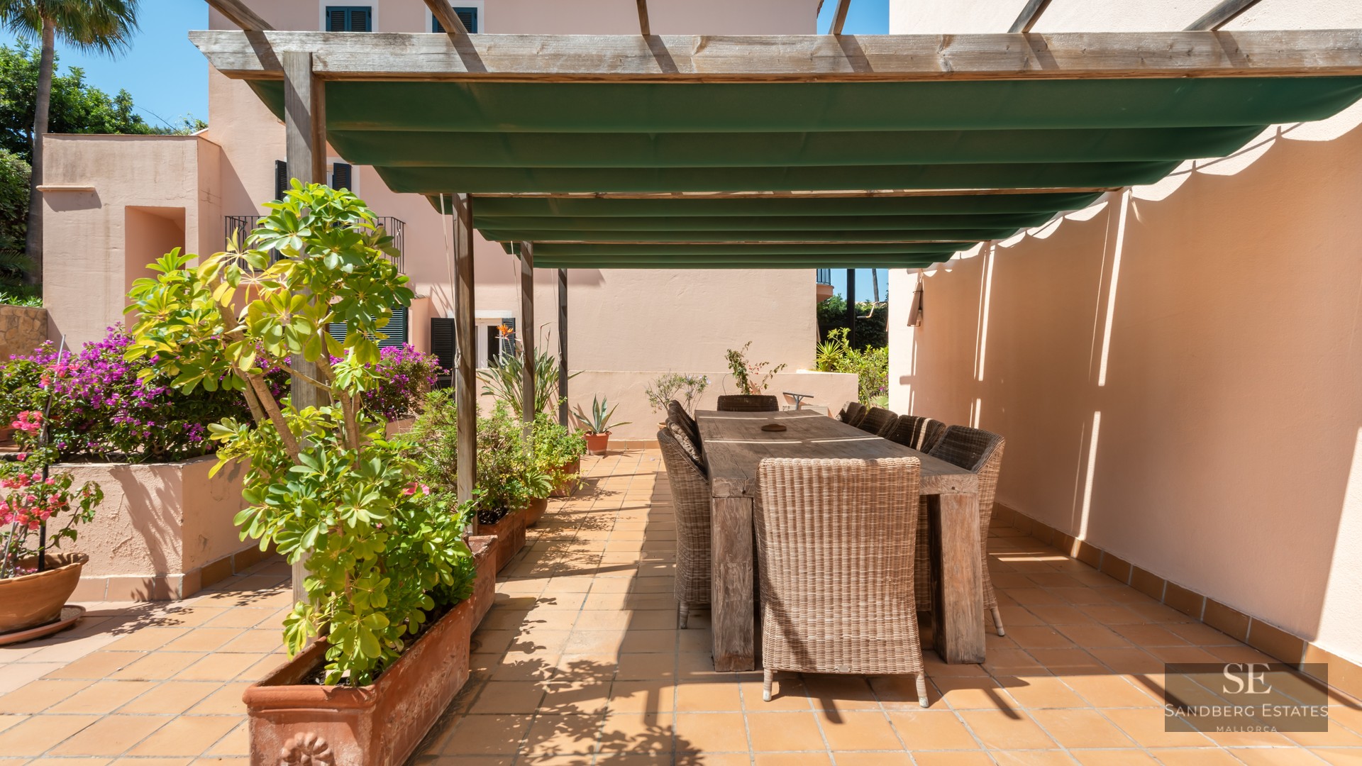 Outdoor dining area on a terracotta terrace with a wooden pergola, green awning, and wicker chairs surrounded by plants.
