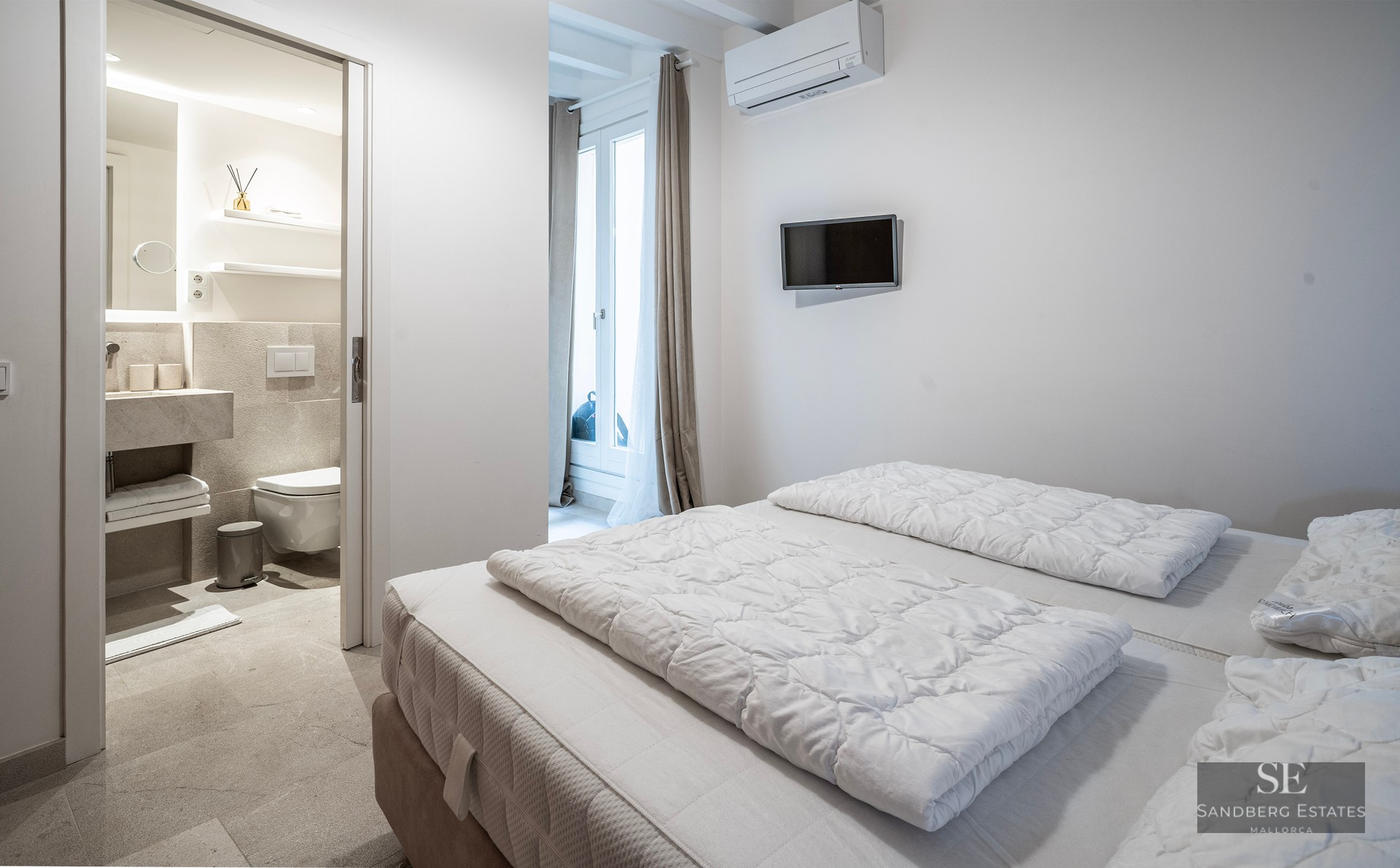Bright modern bedroom with white bedding and open door to a stone-tiled ensuite bathroom.