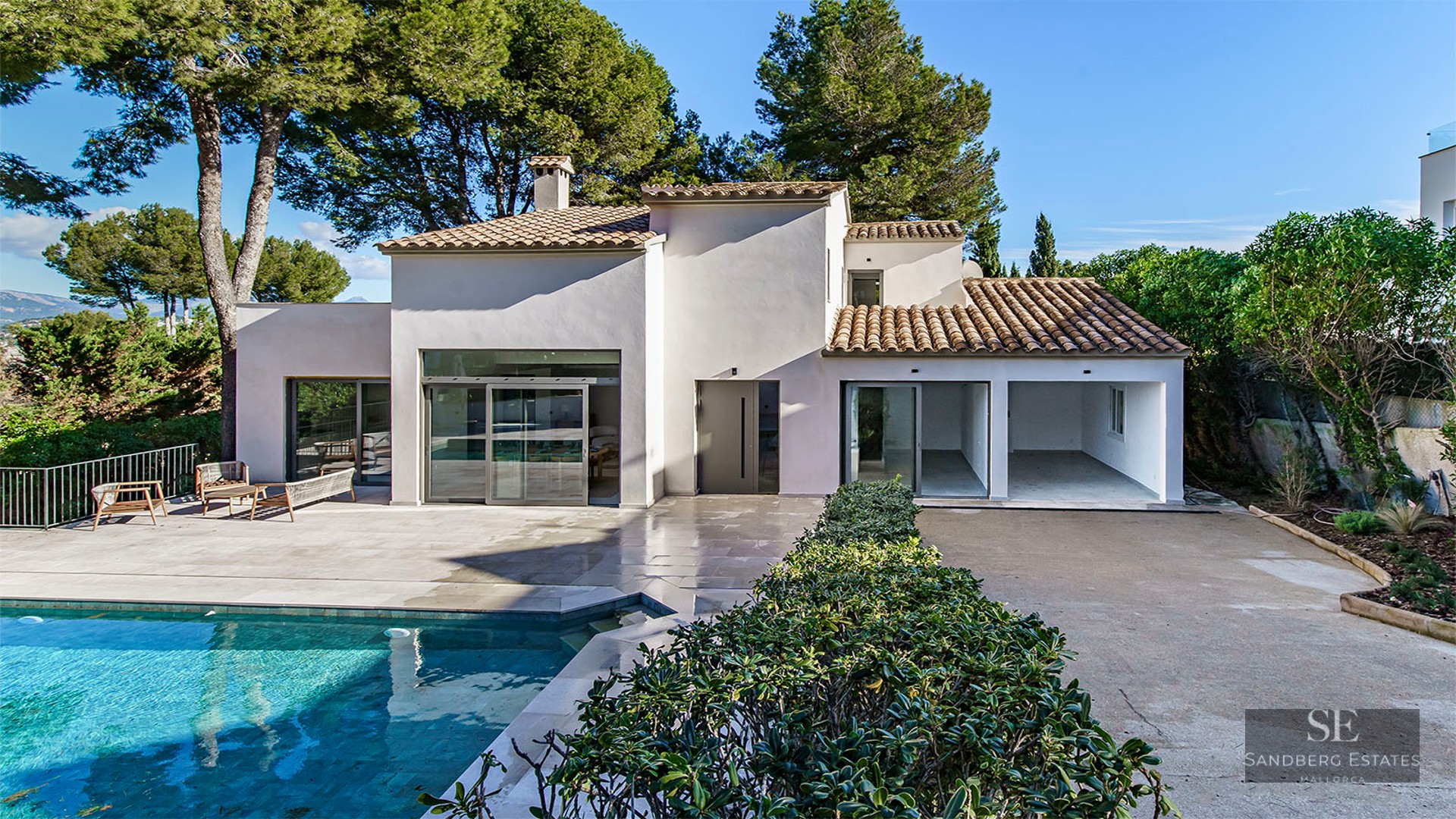 Modern white villa with a turquoise swimming pool, stone terrace, and lush pine trees under a clear blue sky.