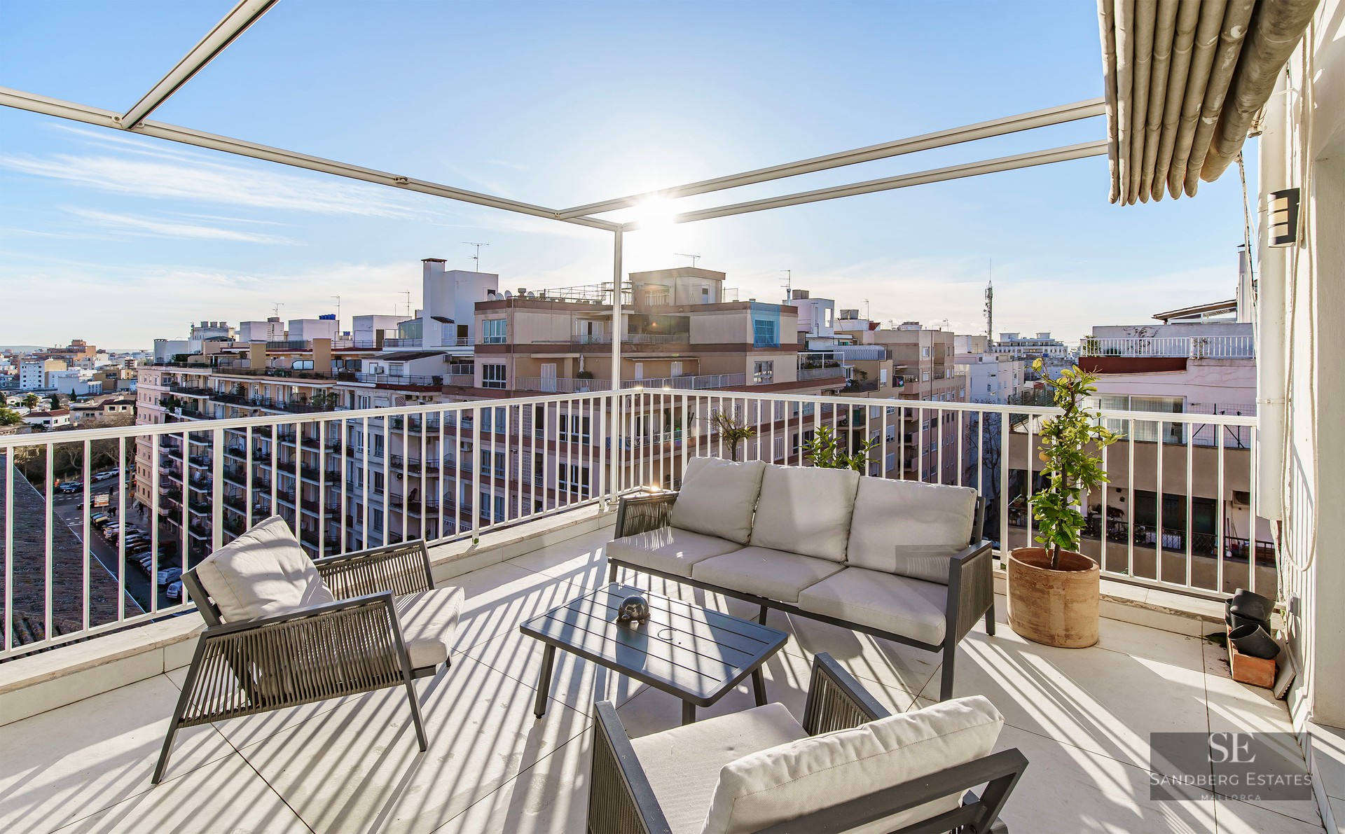 Sun-drenched rooftop terrace with modern grey outdoor furniture, white railings, and an expansive city skyline view.