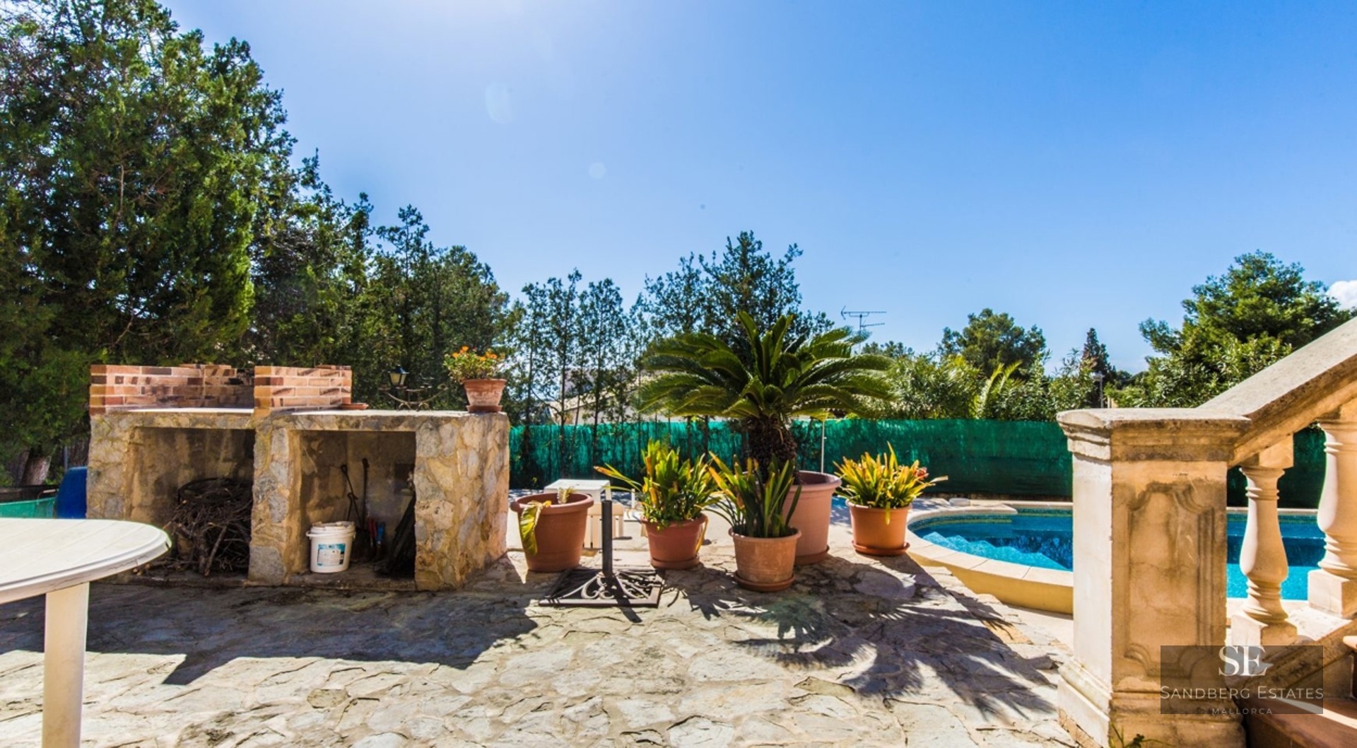 Sun-drenched stone terrace featuring a blue swimming pool, built-in stone barbecue, and lush potted plants under a clear sky.