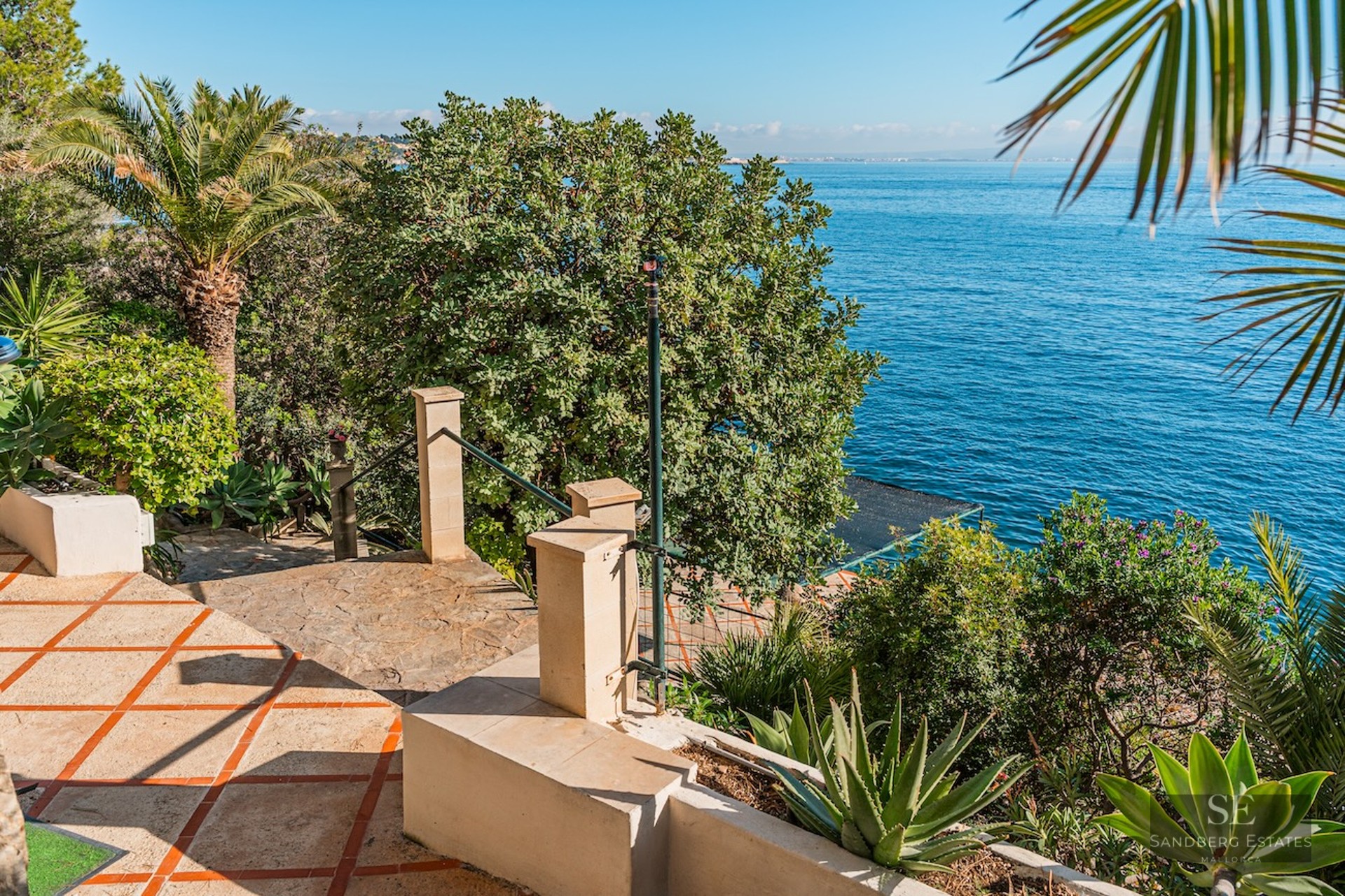 A sunny stone terrace with terracotta tiles leads to a lush garden with palm trees overlooking the blue ocean.