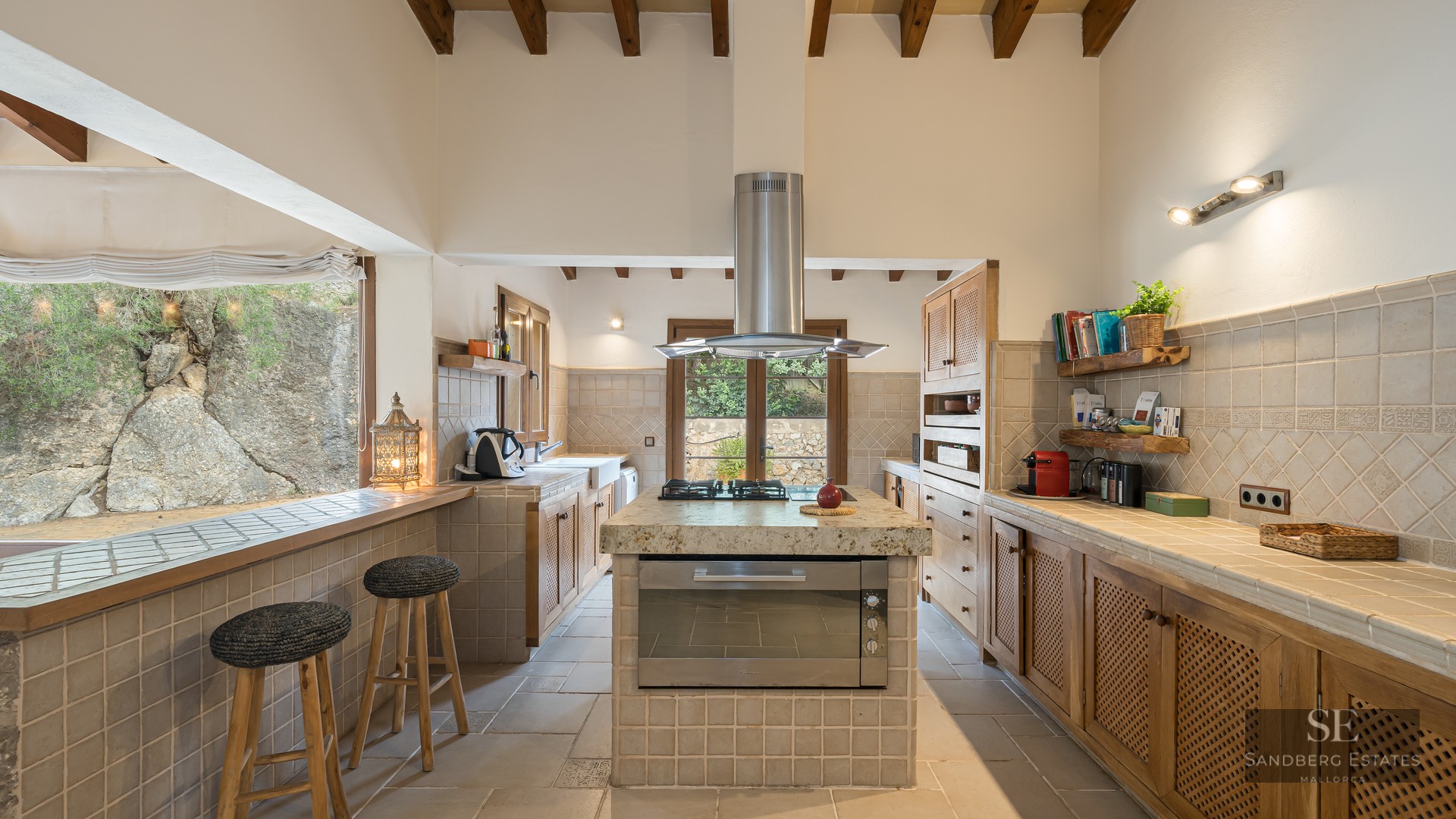 Rustic kitchen featuring wooden beams, a central island, tiled countertops, and bar stools by a window looking out to rocks.