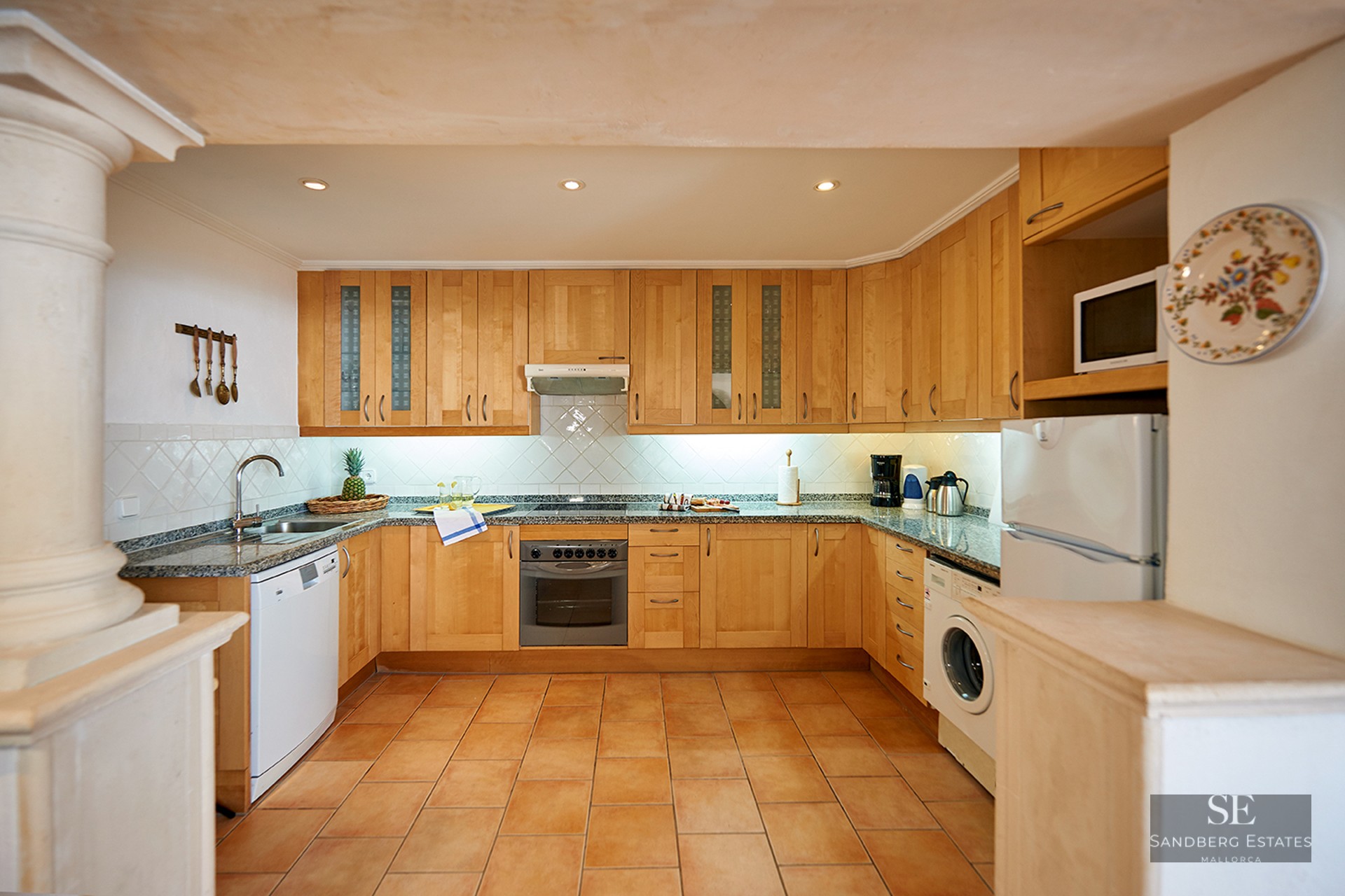 A rustic kitchen with light wood cabinets, granite countertops, terracotta floor tiles, and appliances.
