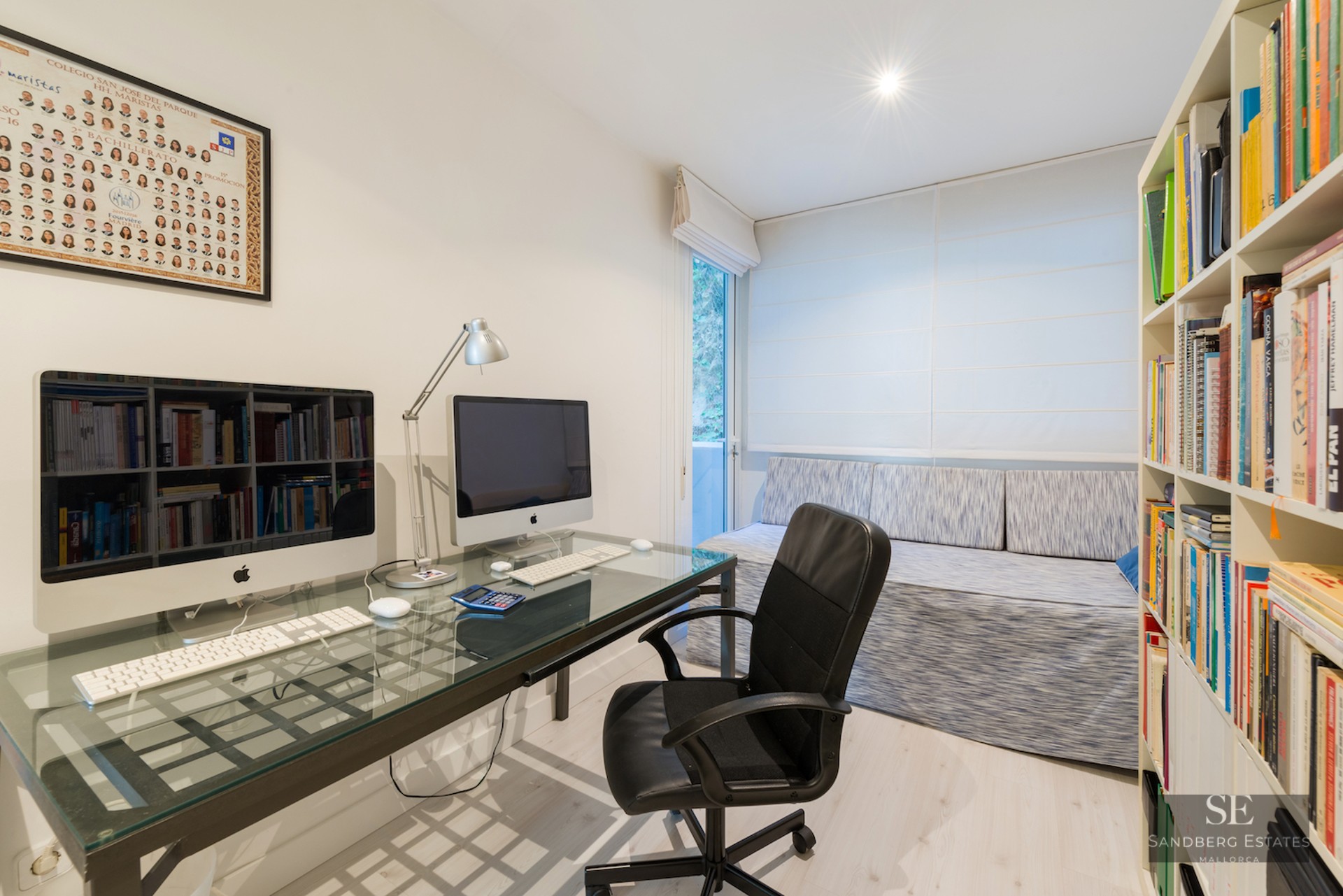 A bright home office featuring a glass desk with two computers, a black ergonomic chair, and floor-to-ceiling bookshelves.