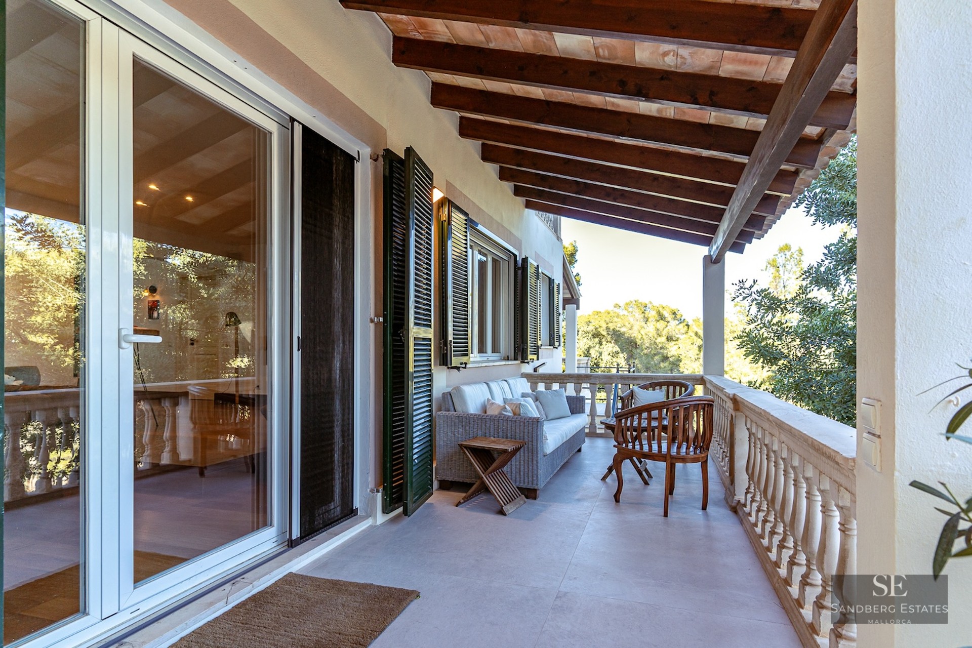 Covered terrace with wooden ceiling beams, wicker sofa, stone balustrade, and a view of lush greenery.