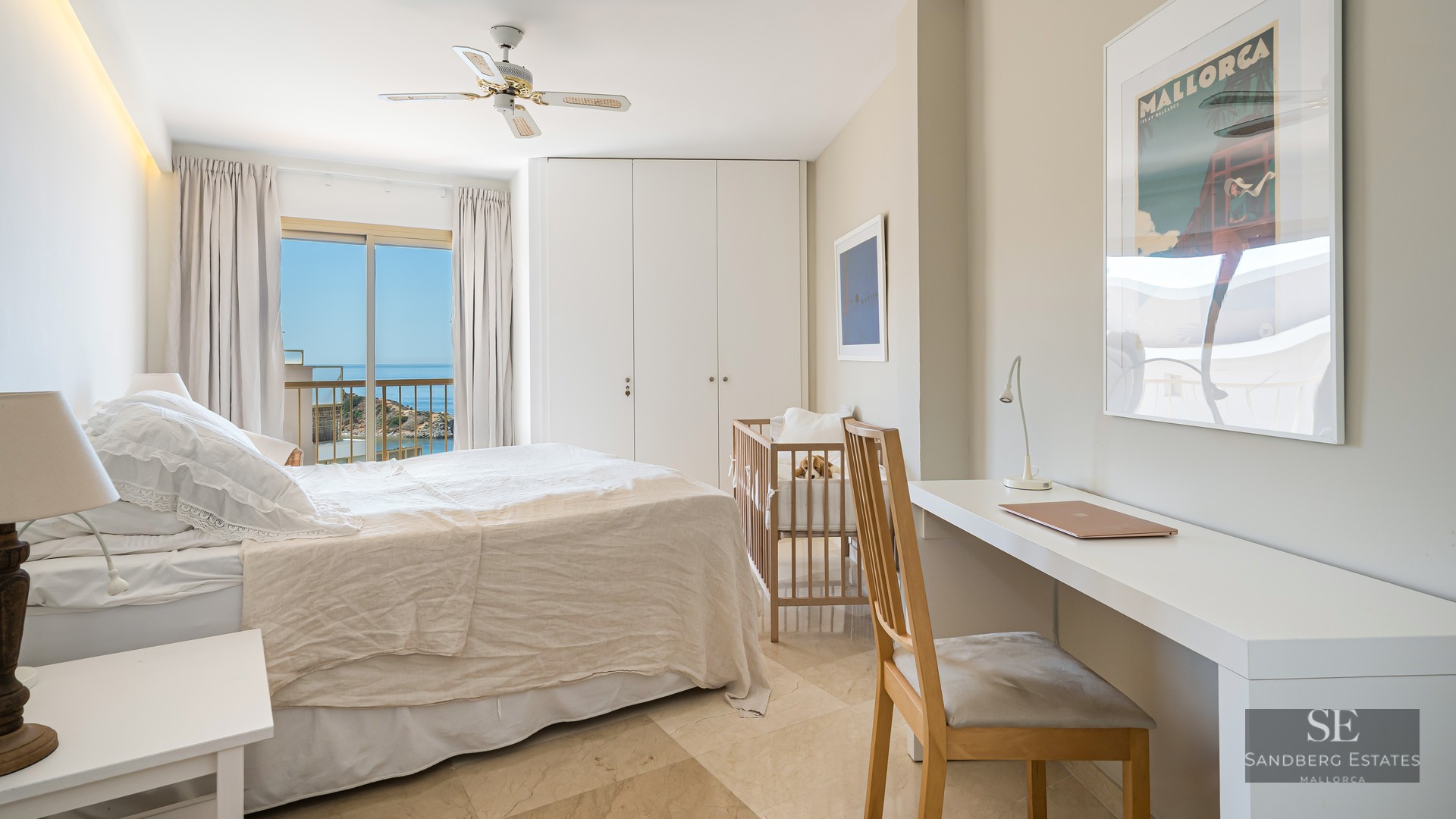 Modern bedroom featuring a white bed, baby crib, desk, and balcony with panoramic sea views.