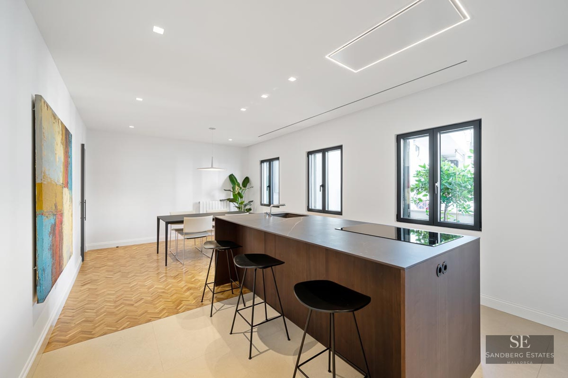 Minimalist kitchen featuring a dark wood island, black bar stools, herringbone wood flooring, and abstract wall art.