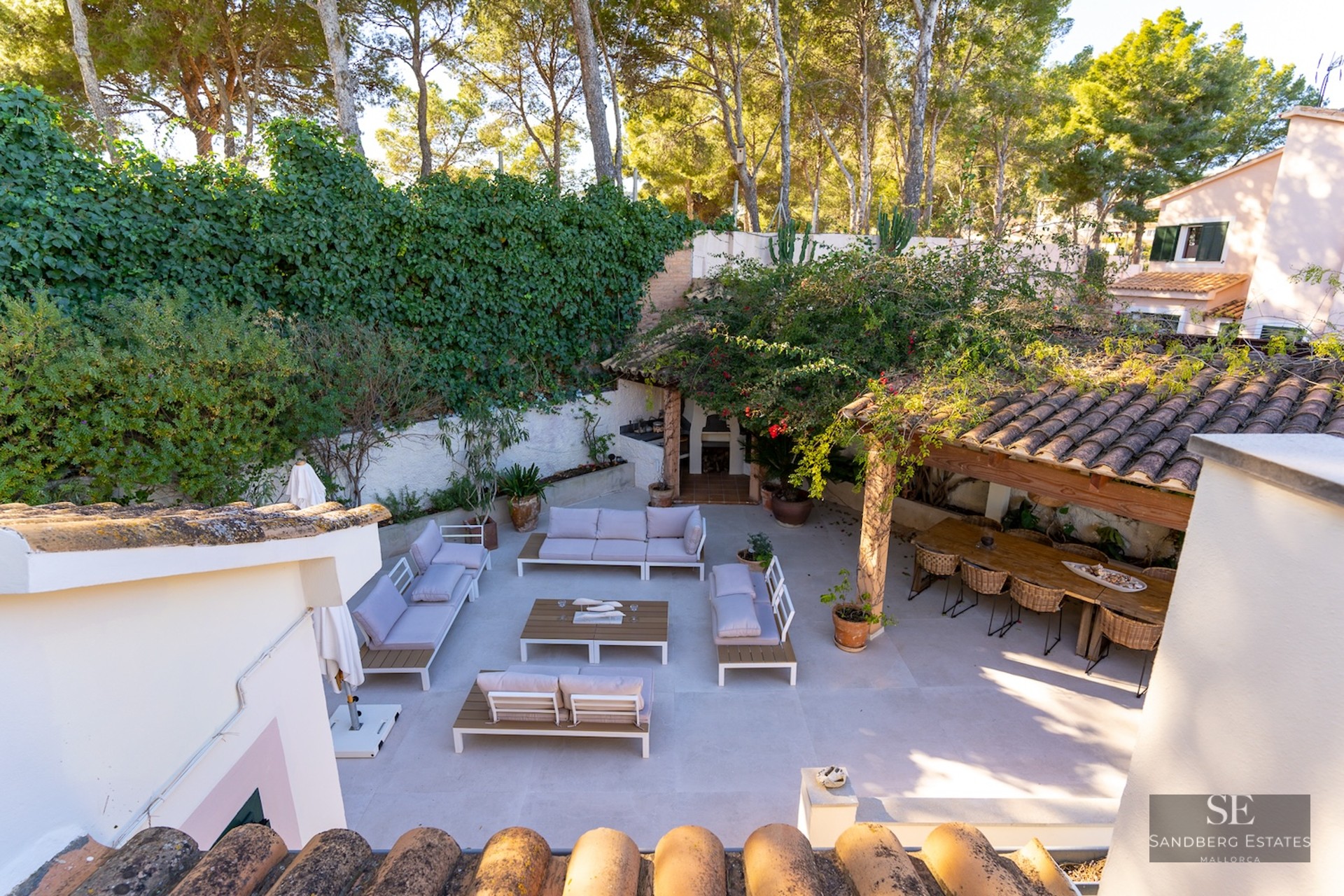 Elevated view of a spacious paved terrace with white lounge sofas, a large wooden dining table, and lush green walls.