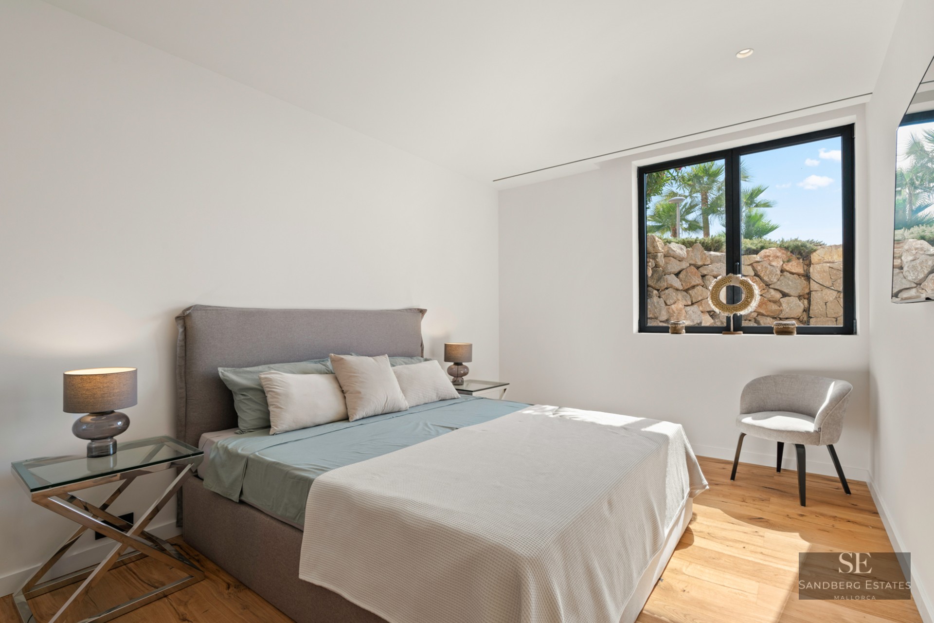 Bright modern bedroom with a gray upholstered bed, glass nightstands, wooden floors, and a large window.