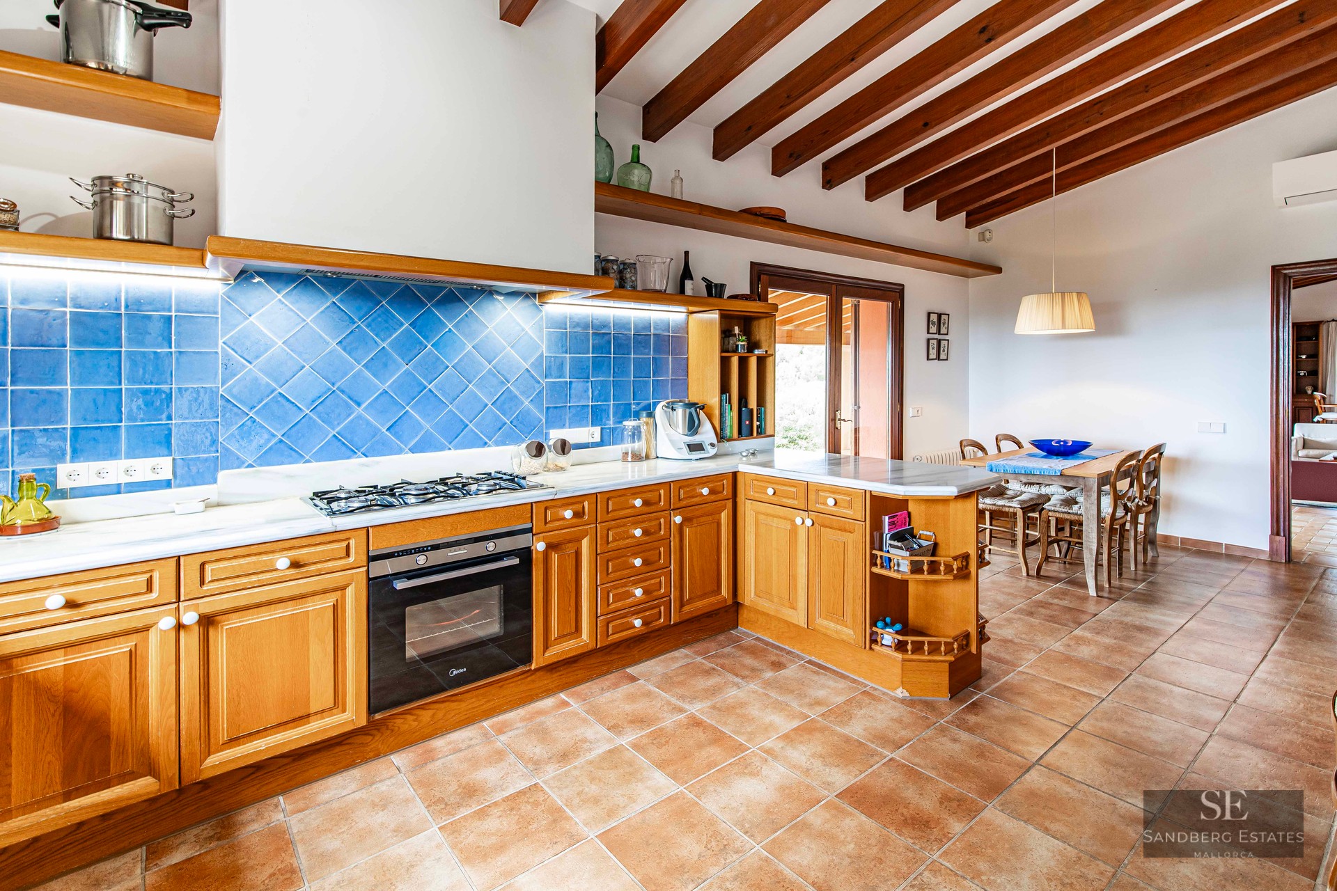Rustic kitchen featuring wooden cabinets, blue tiled backsplash, terracotta floors, and exposed ceiling beams.