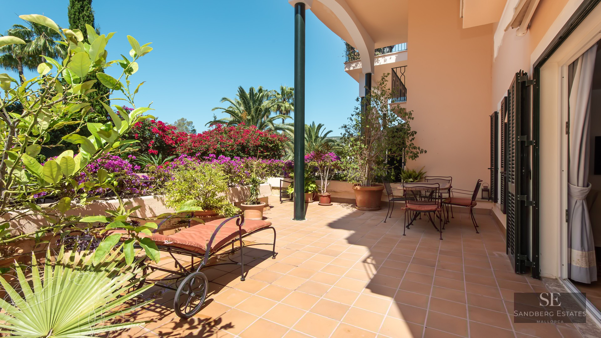Terracotta terrace with a sun lounger and dining area surrounded by bougainvillea and palm trees under a blue sky.