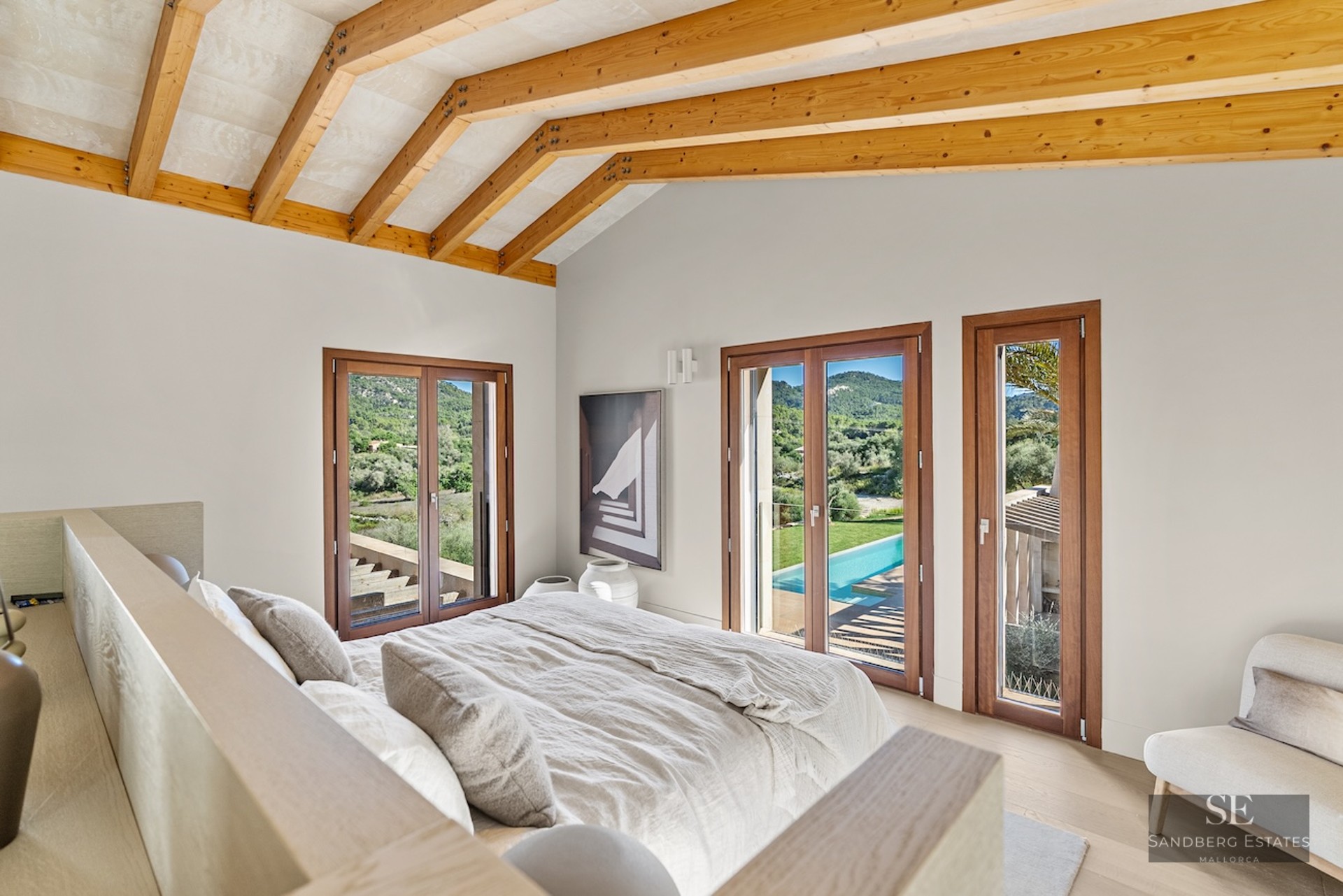 Modern bedroom with vaulted wood beam ceiling, bed with beige linens, and glass doors overlooking a pool and green hills.