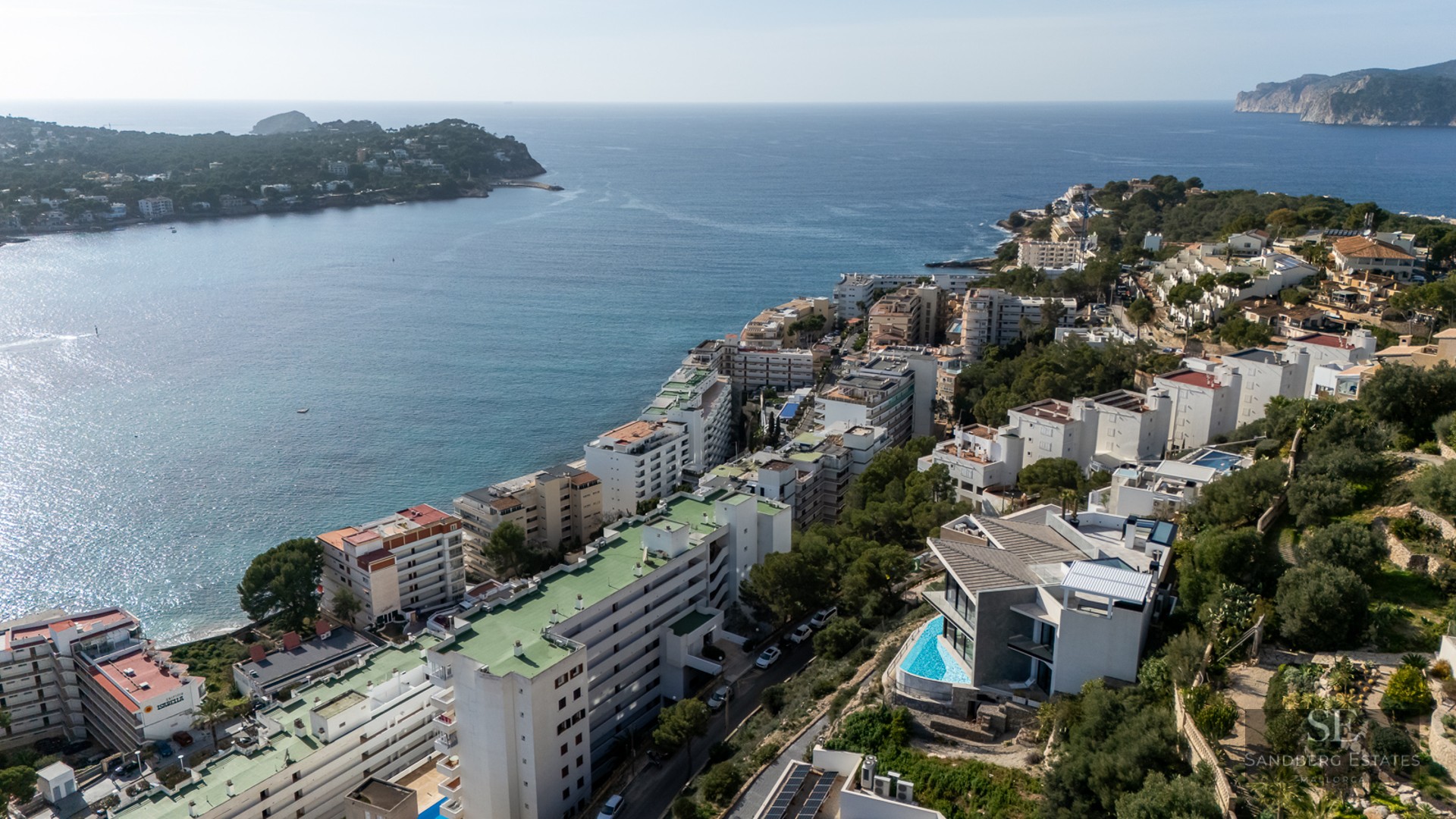 Aerial view of white coastal buildings lining a blue bay and green hills.