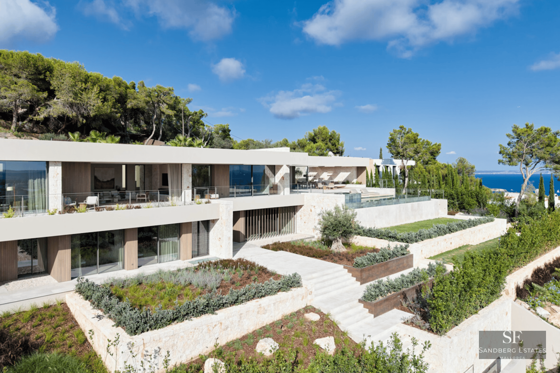 Modern two-story luxury villa with stone facades, glass balconies, and tiered Mediterranean gardens overlooking the sea.