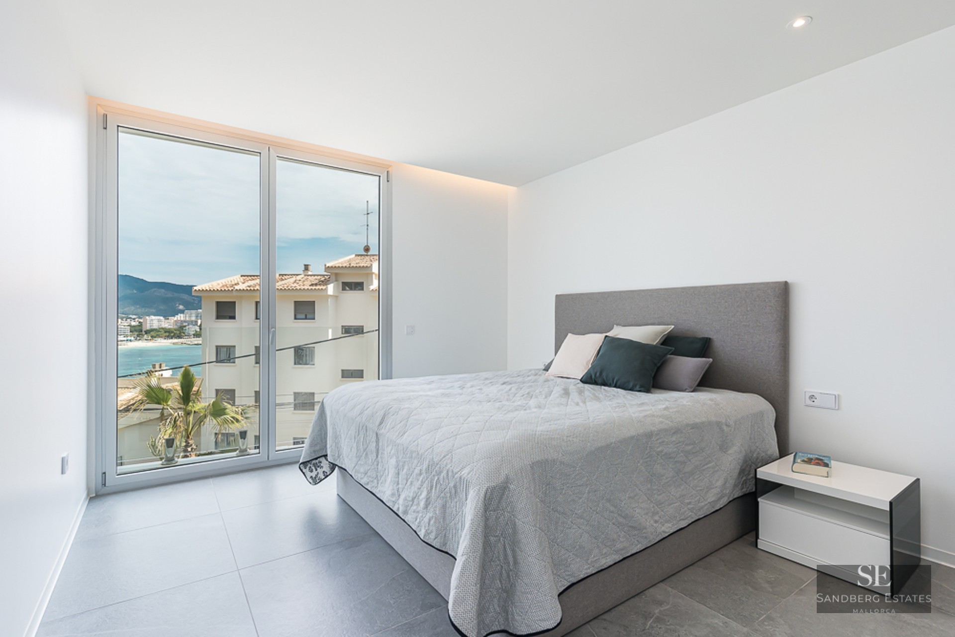 Bright modern bedroom with a large window overlooking a coastal town and sea, featuring a grey bed and tiled floor.