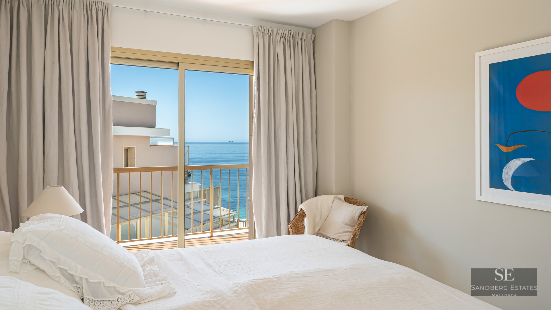Sunlit bedroom featuring a white bed, wicker chair, and sliding glass doors opening to a sea view balcony.