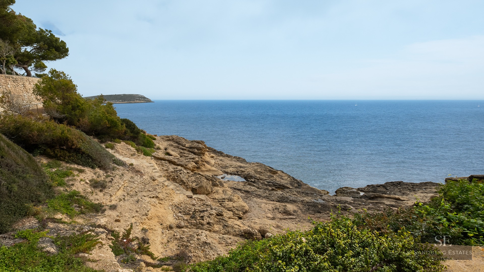 Rocky coastline meeting the blue sea with green Mediterranean shrubs in the foreground.